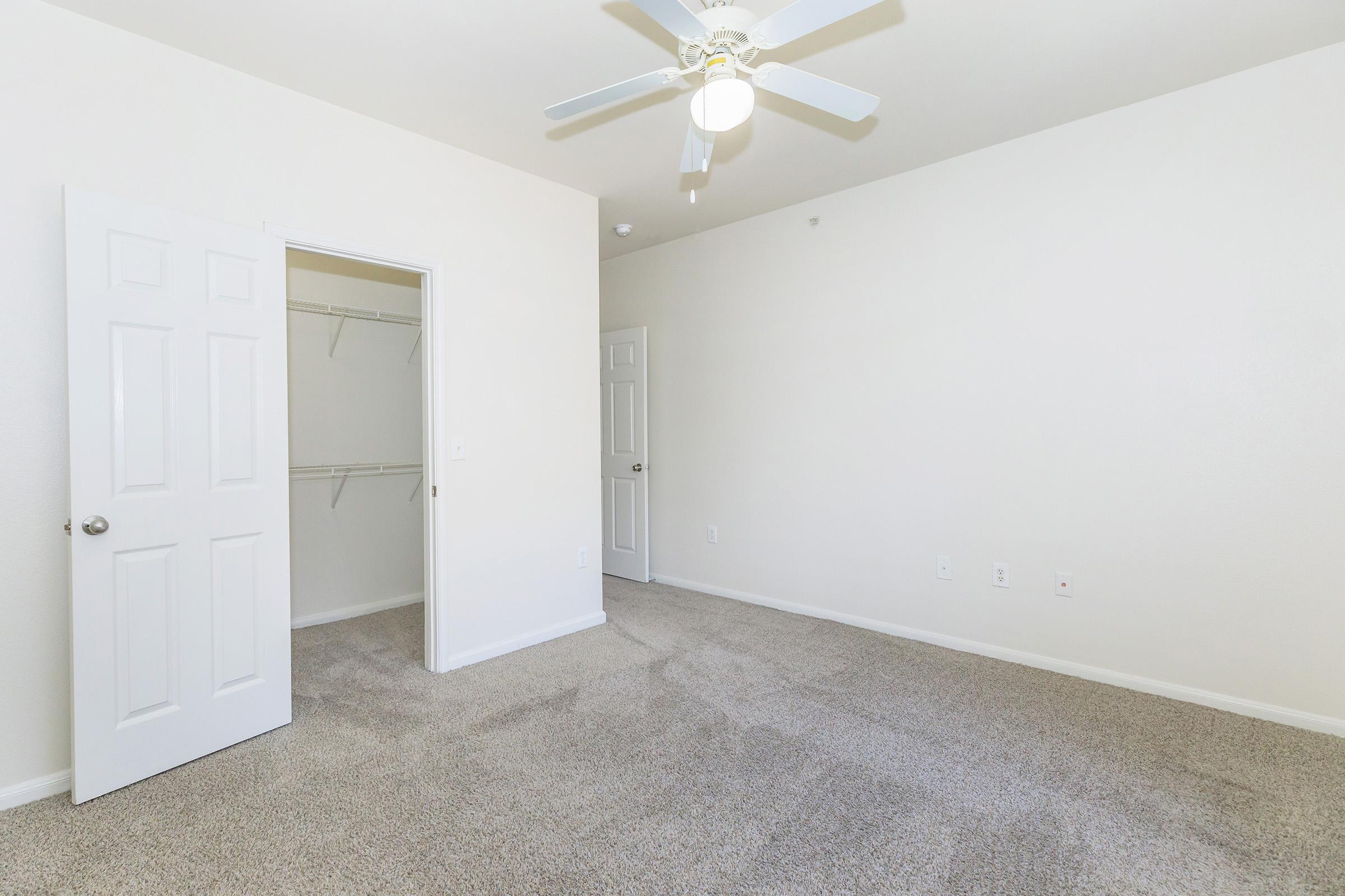 A neutral-colored room with light beige carpeting, featuring a ceiling fan and white walls. There are two doors: one leading to a closet and another to an adjoining room. The space is bright and empty, providing a clean and simple aesthetic suitable for various furnishings.