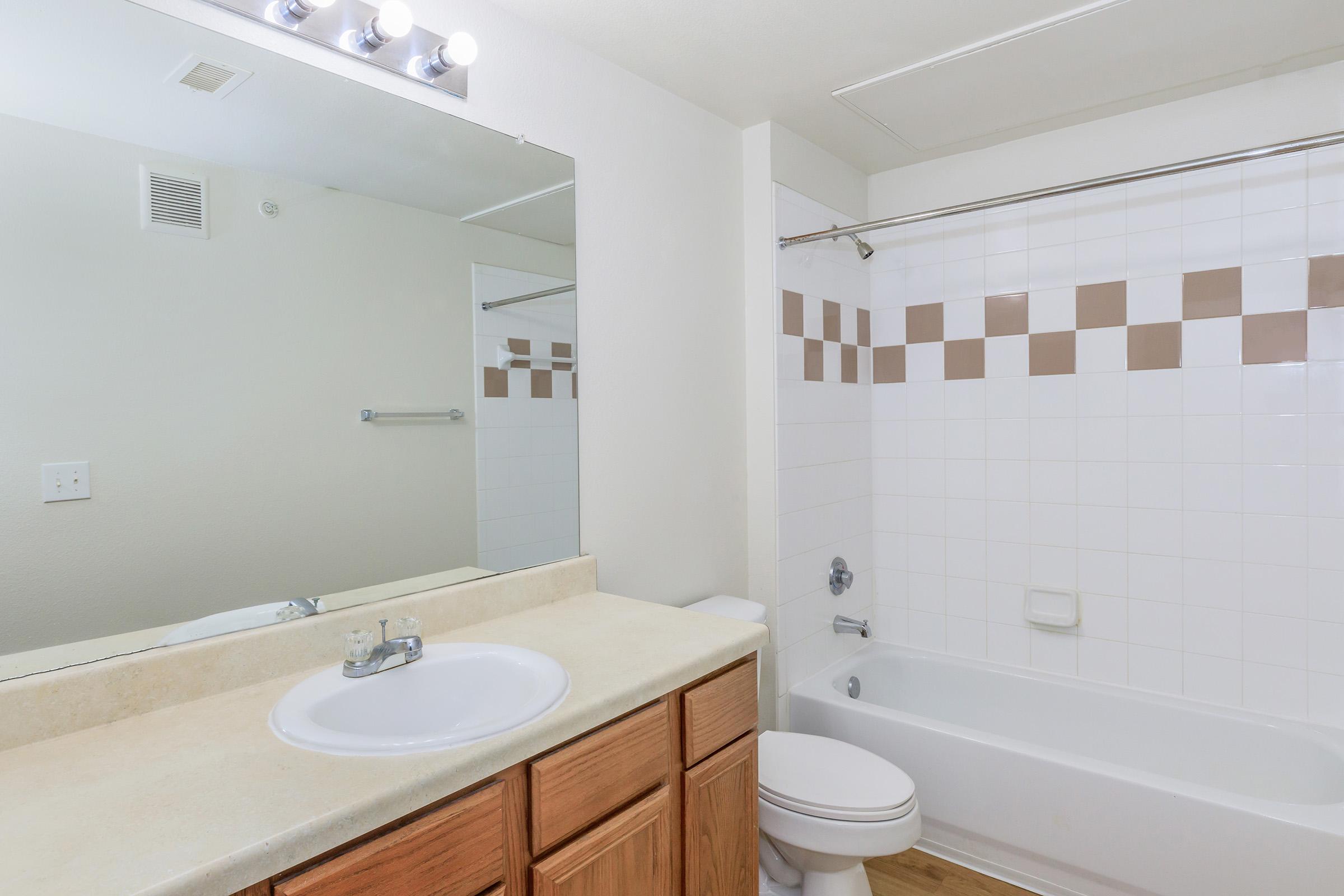 A clean, modern bathroom featuring a white bathtub with a shower, beige and brown tiled accents, a countertop with a sink, and a large mirror above. The walls are light-colored, and the flooring appears to be wood laminate. The overall atmosphere is bright and tidy.