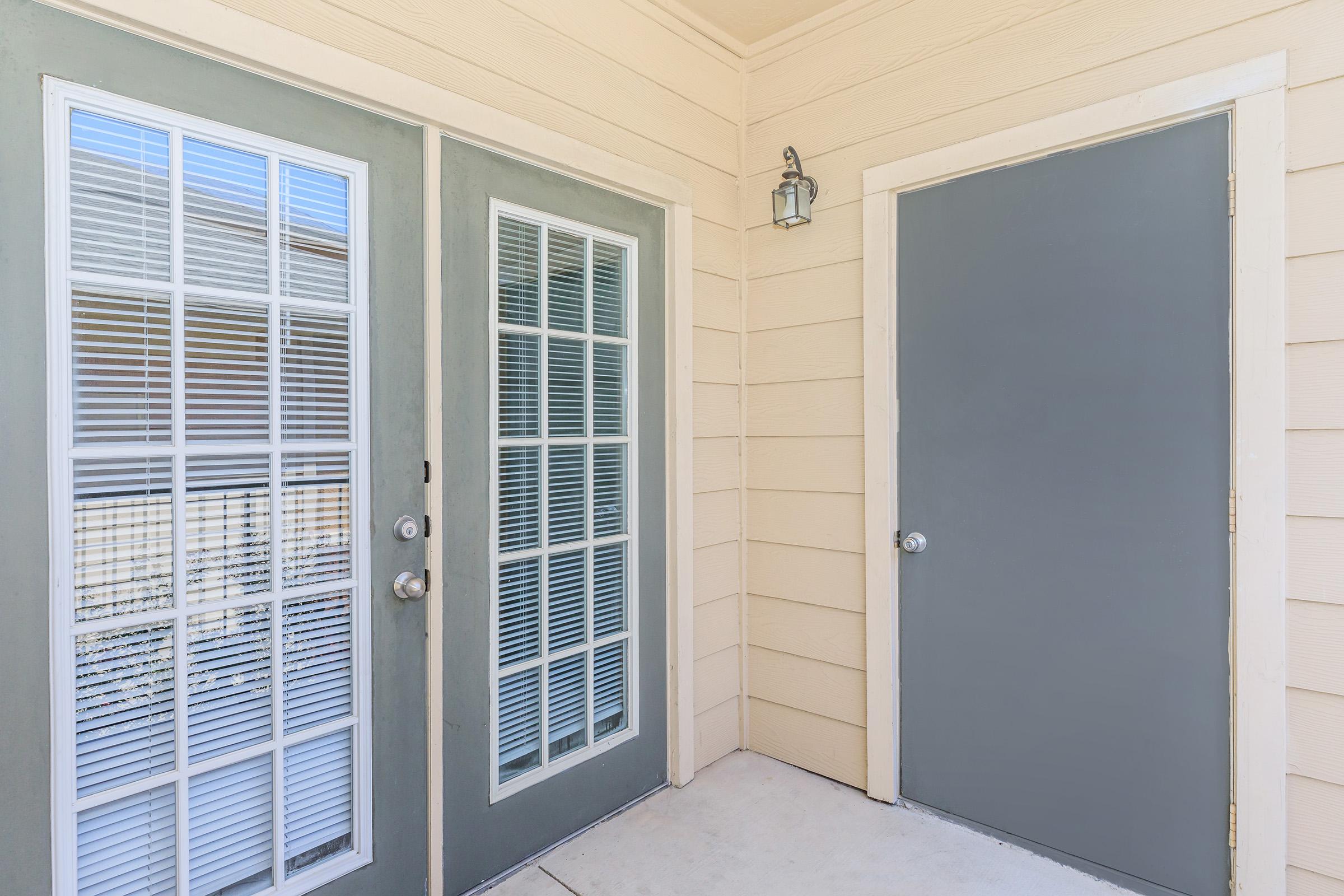 An entrance area featuring two sets of doors. The left door has multiple square windows with white blinds, while the right door is solid gray. The walls are light-colored with a smooth finish, and a small outdoor light fixture is mounted above the doors. The floor is concrete.