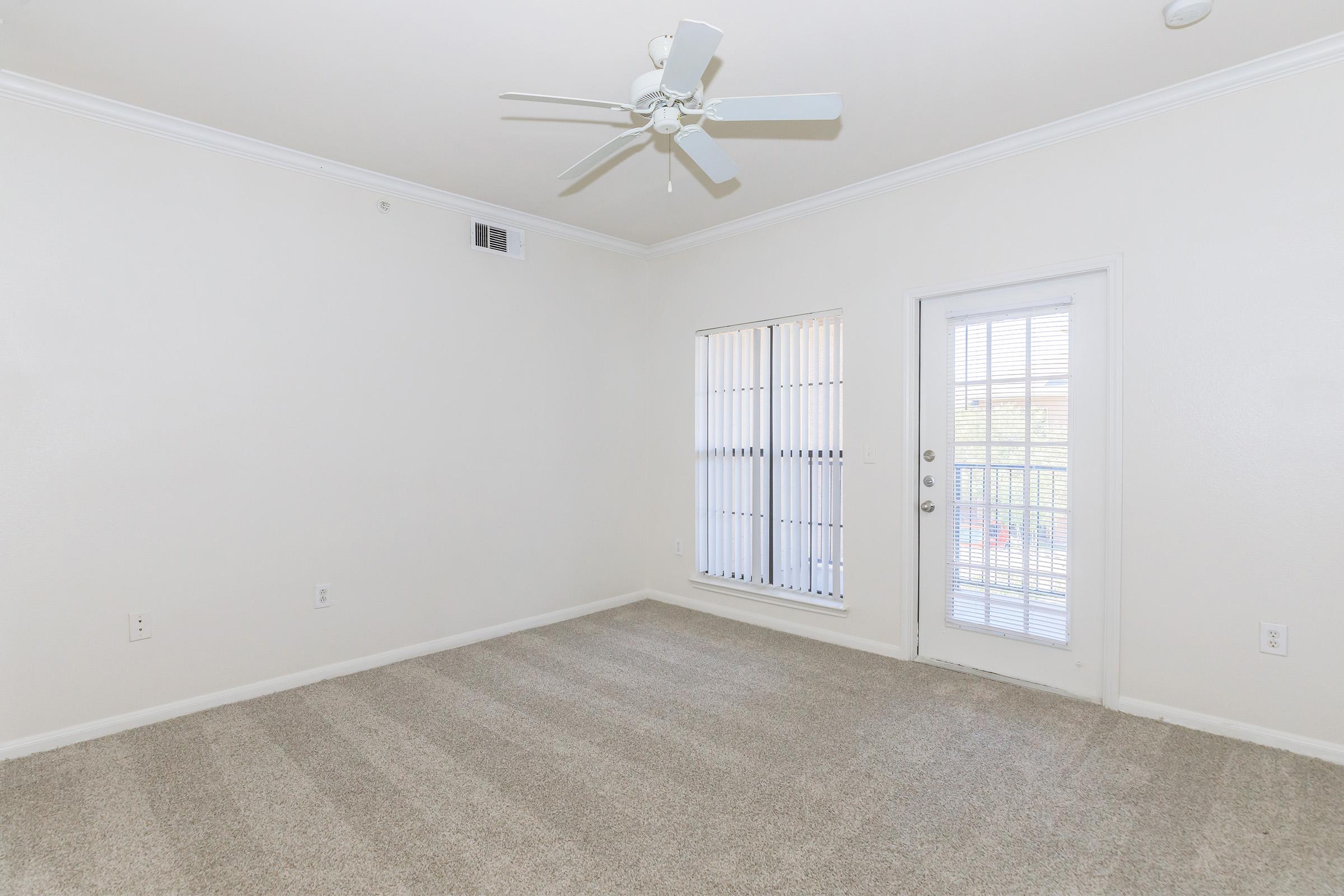 A spacious, empty room with light beige walls and carpet. It features a ceiling fan, a large window with vertical blinds, and a door leading to a balcony. The room is well-lit and designed for versatility, suitable for various furnishing options.