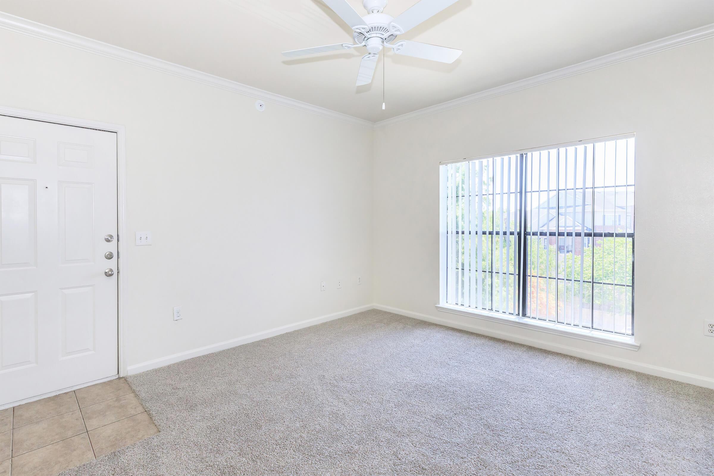 Interior of a room featuring light-colored walls, a ceiling fan, and a window with blinds. The floor is carpeted and there is a tiled entryway. The space appears empty and is well-lit by natural light coming through the window.
