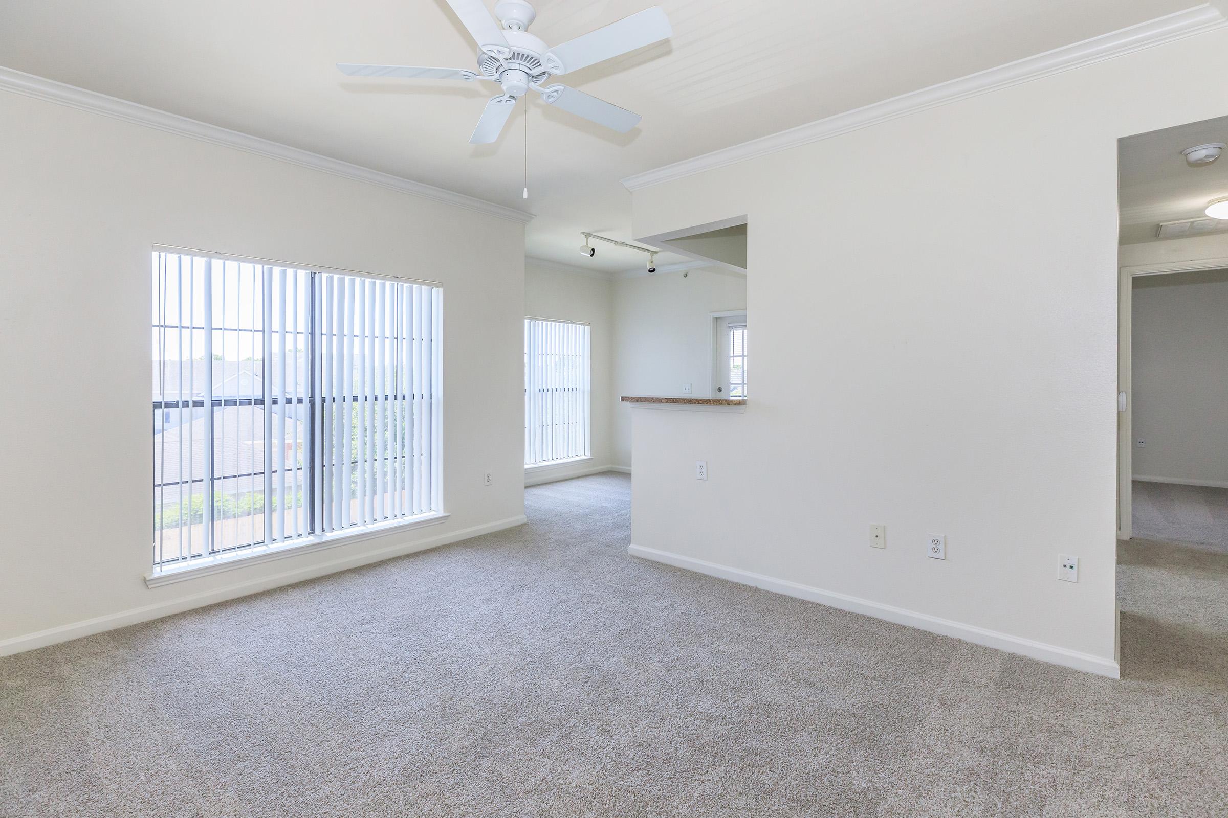 Bright and spacious living area with light-colored walls and carpet, featuring a ceiling fan. Large windows with vertical blinds allow natural light. A small bar separates the living space from the kitchen area. The layout is open and inviting, showcasing a clean and modern design.