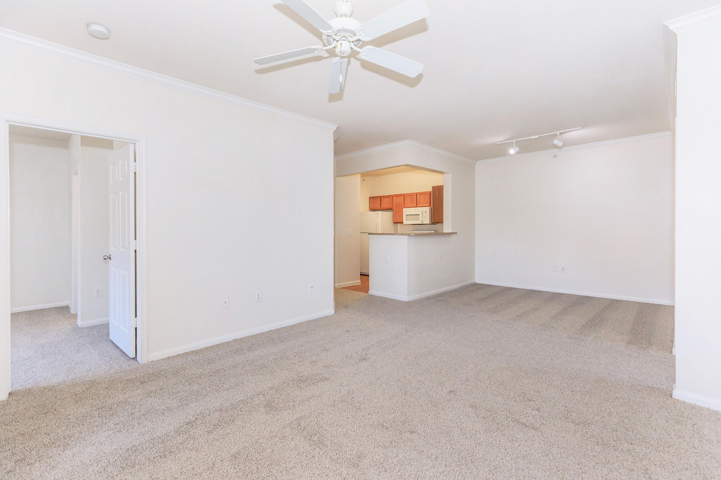 An empty, well-lit living space featuring light beige walls, cream carpeting, and a ceiling fan. The room has an open layout with a doorway on the left leading to another room, and a kitchen visible in the background with wooden cabinets and appliances. Ideal for a cozy, modern apartment setting.