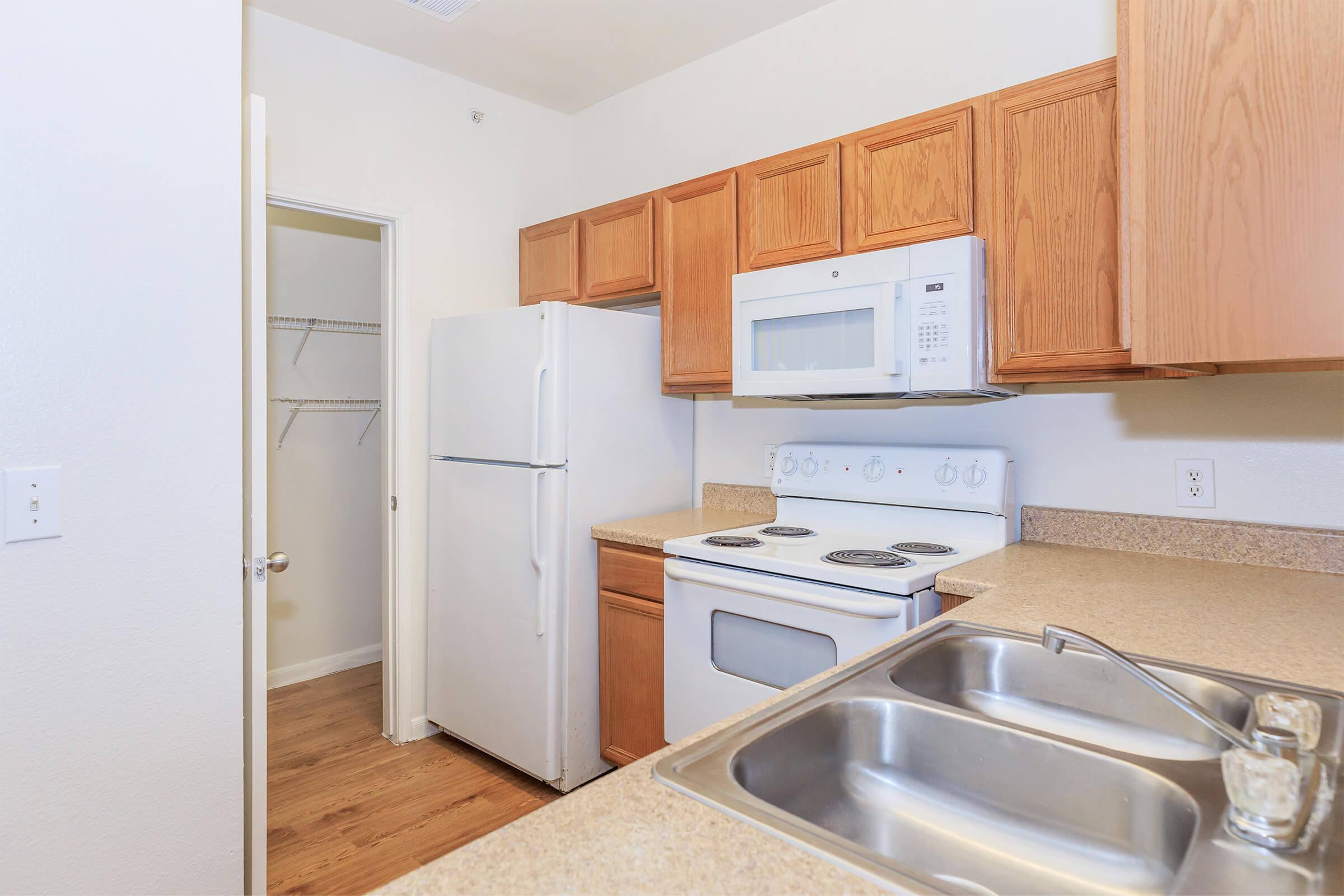 A view of a small kitchen featuring wooden cabinets, a white refrigerator, a microwave above a white stove, and a countertop with a double sink. A doorway leads to a pantry or closet, and the floor appears to be hardwood. The walls are painted in a light color.