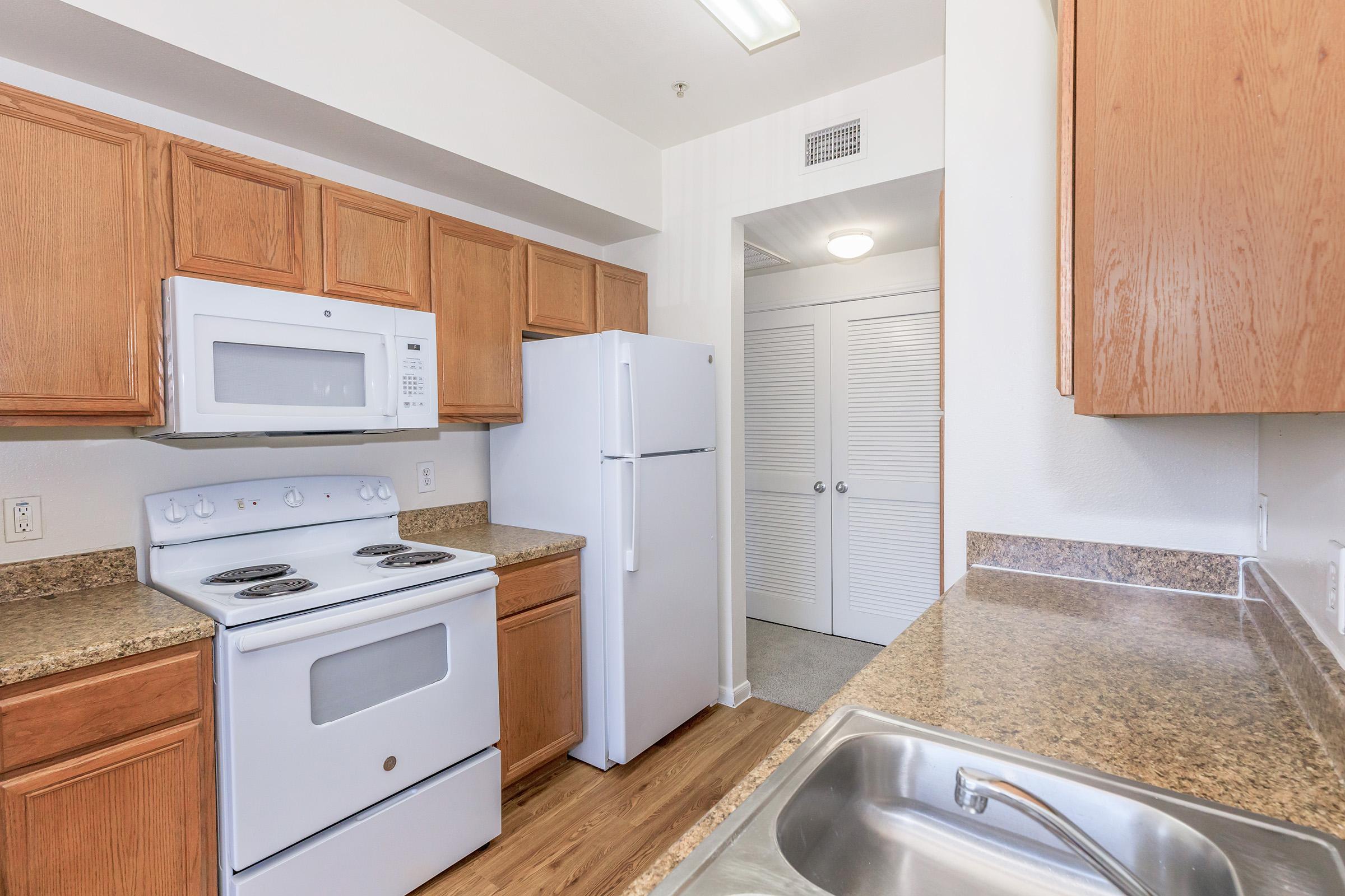 A modern kitchen featuring wooden cabinets, a white microwave, and refrigerator. The countertop is speckled, and there's a stainless steel sink with a faucet. In the background, a door leads to a room with white bi-fold closet doors. The overall color scheme is light and inviting.