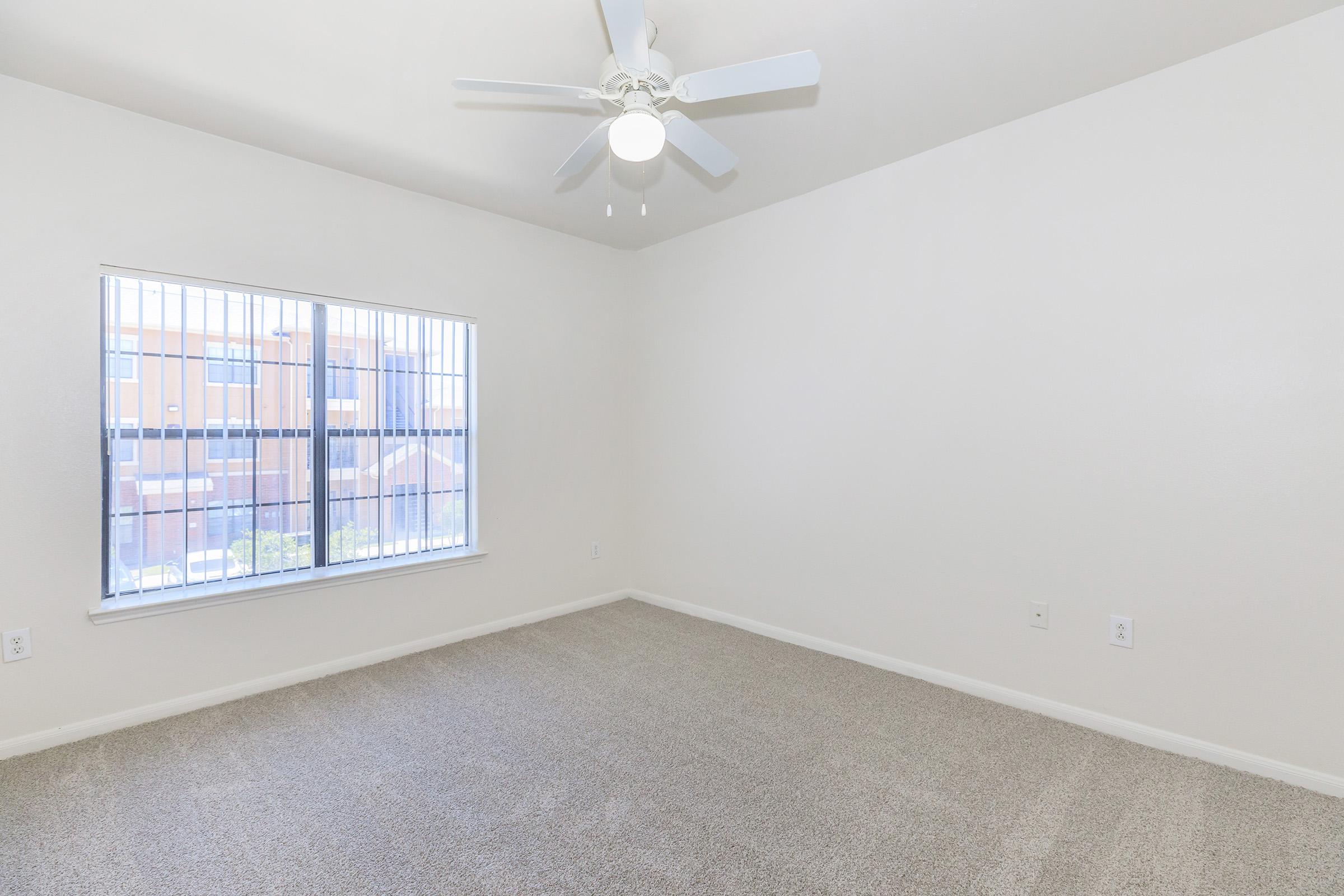 A clean, empty bedroom with light beige walls and carpet. There's a ceiling fan with white blades, and a large window with vertical blinds allowing natural light to enter. The view outside the window shows a colorful building in the background. The room appears bright and spacious.