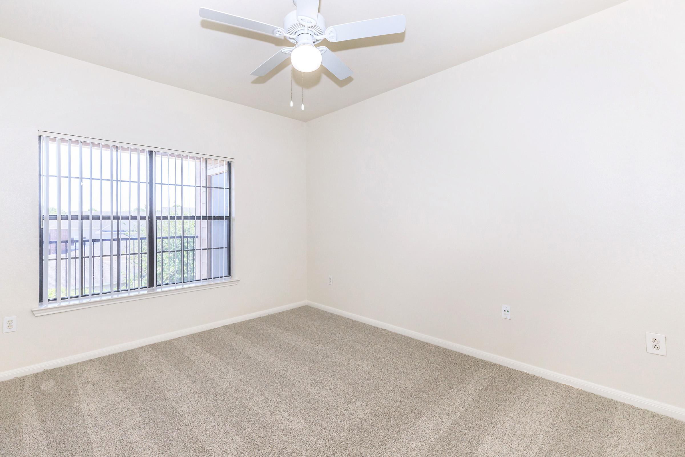 Empty room with light-colored walls and carpeted floor. A ceiling fan is installed in the center, and a window with vertical blinds is visible, providing natural light. The overall space appears clean and well-lit, suitable for various uses.
