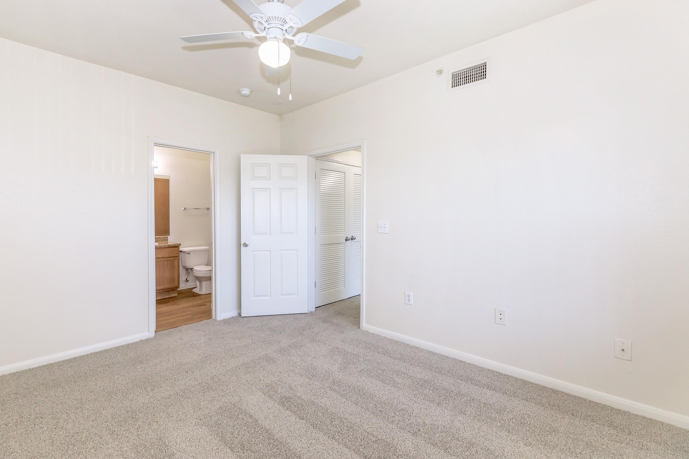 A spacious, well-lit room with light beige walls and plush carpeting. A ceiling fan hangs from the ceiling. One door leads to a bathroom, visible just off to the side, while another door leads to an adjacent room. The overall feel is modern and inviting, suitable for a bedroom or living space.