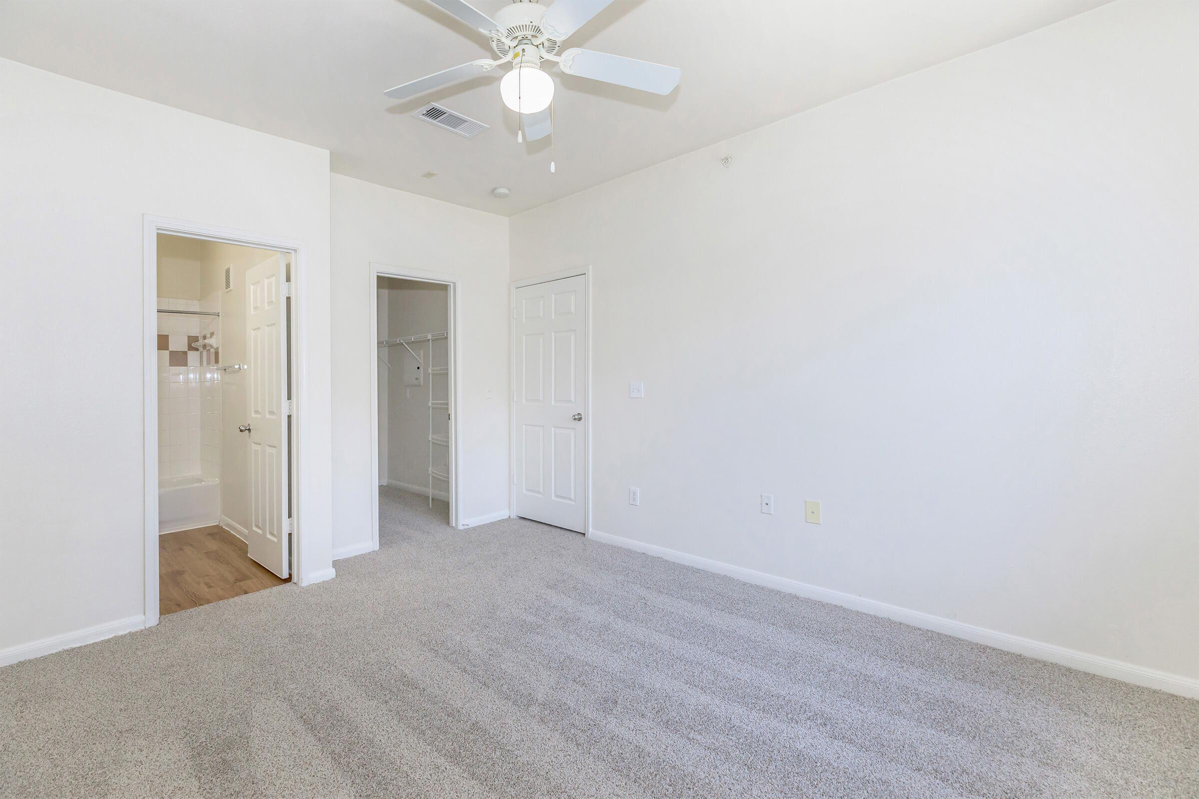 A spacious, empty bedroom featuring light-colored walls and carpet. A ceiling fan is installed, and there are two doors: one leading to a bathroom and the other to a closet. The room is well-lit with natural light and has a neutral, clean aesthetic.