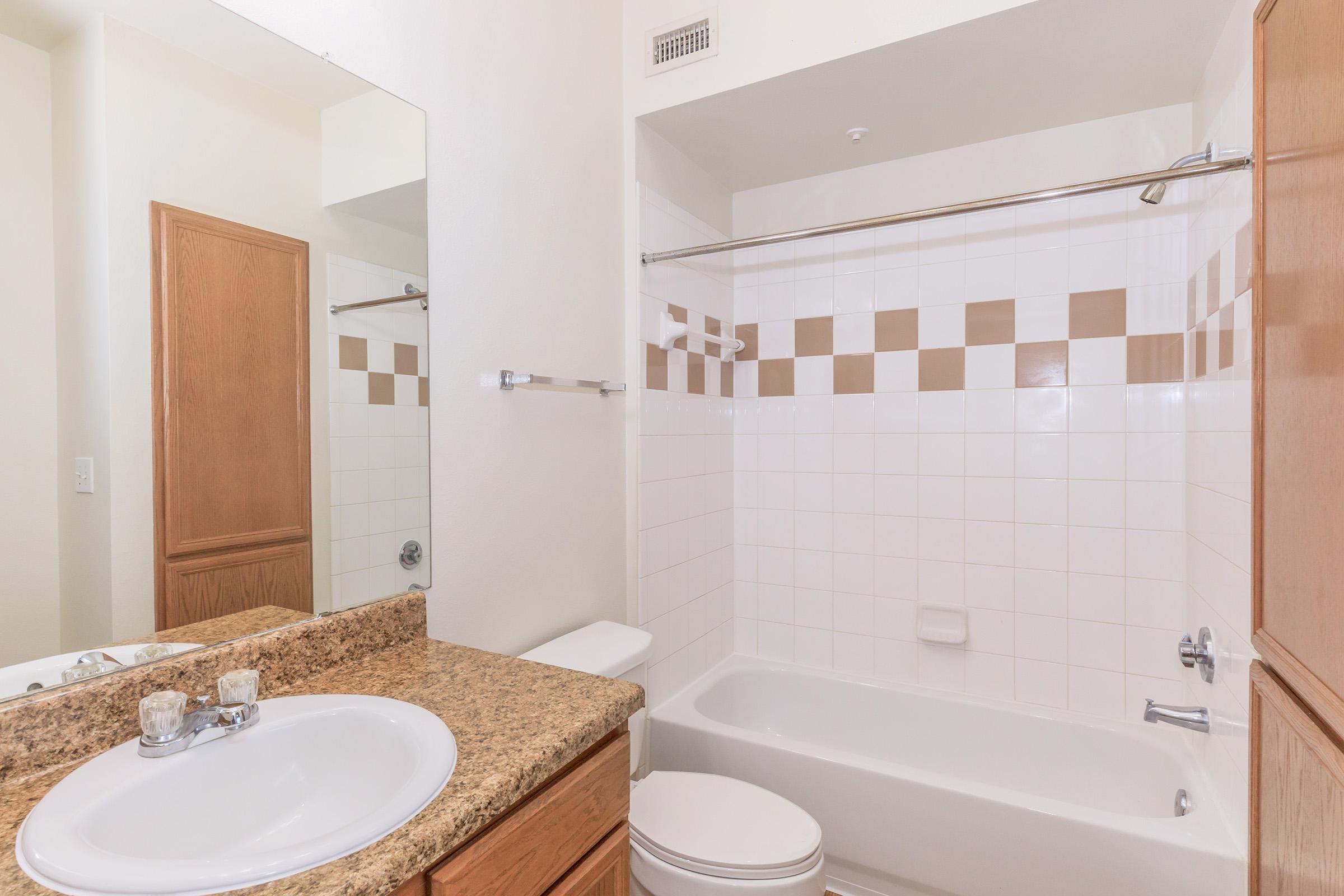 A clean and modern bathroom featuring a bathtub, toilet, and a sink with a granite countertop. The wall has white tiles with a brown checkerboard design. A large mirror reflects part of the room, and the overall decor is simple and contemporary, enhancing the bright and spacious feel.