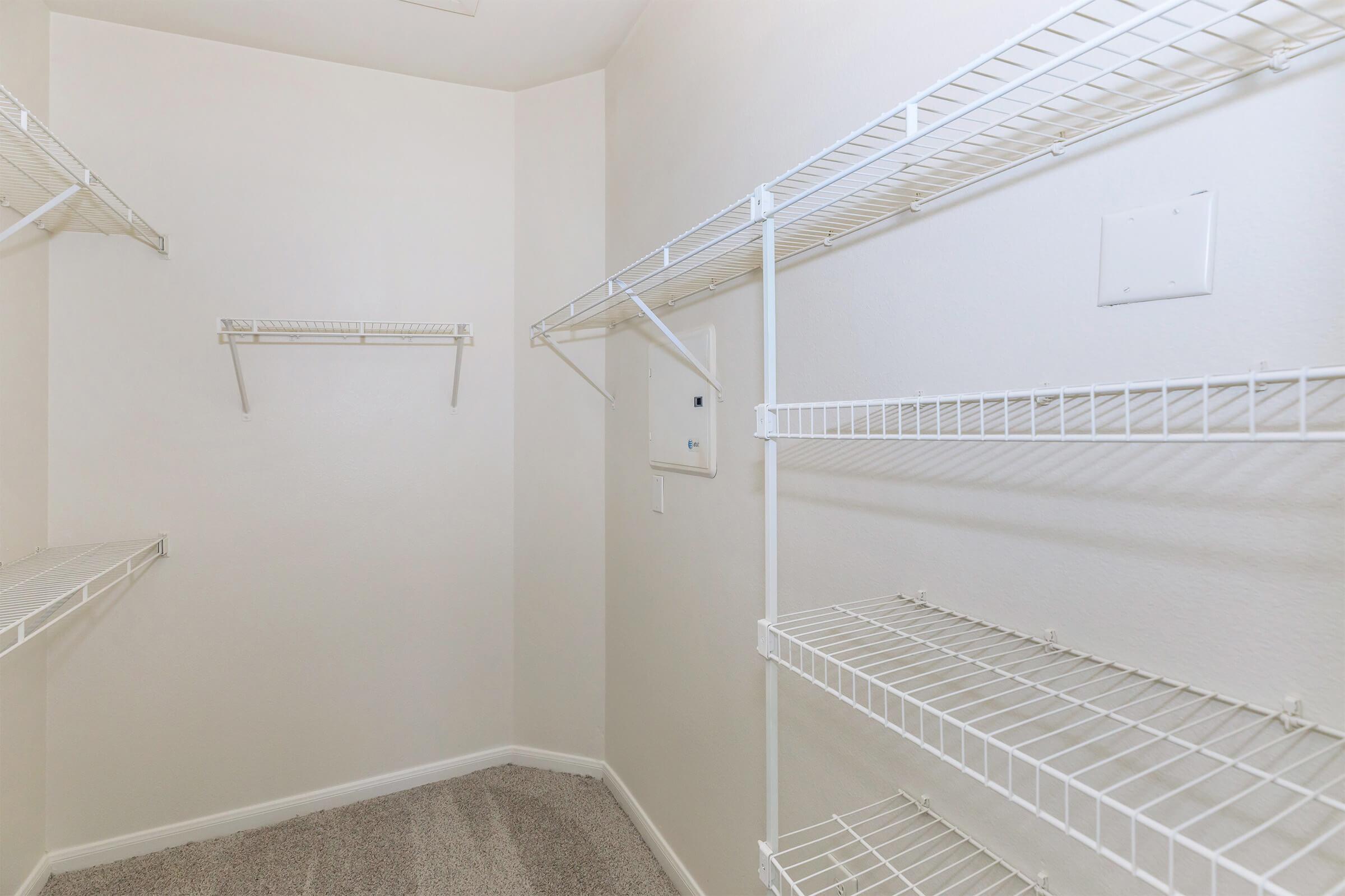 A spacious walk-in closet with white wire shelving units mounted on light-colored walls. The floor is carpeted, and there is a small white panel on one wall, adding to the closet's organized design.