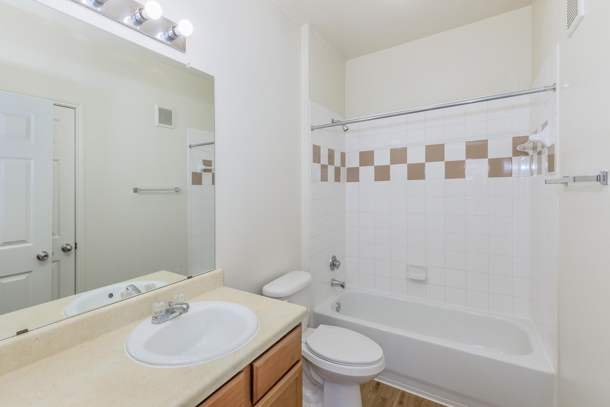 A bright and clean bathroom featuring a bathtub with a shower, a mirror above the sink, and beige and white tiled walls. The sink is circular and made of light-colored stone, while the cabinetry below is wooden. The room has a neutral color scheme and good lighting.
