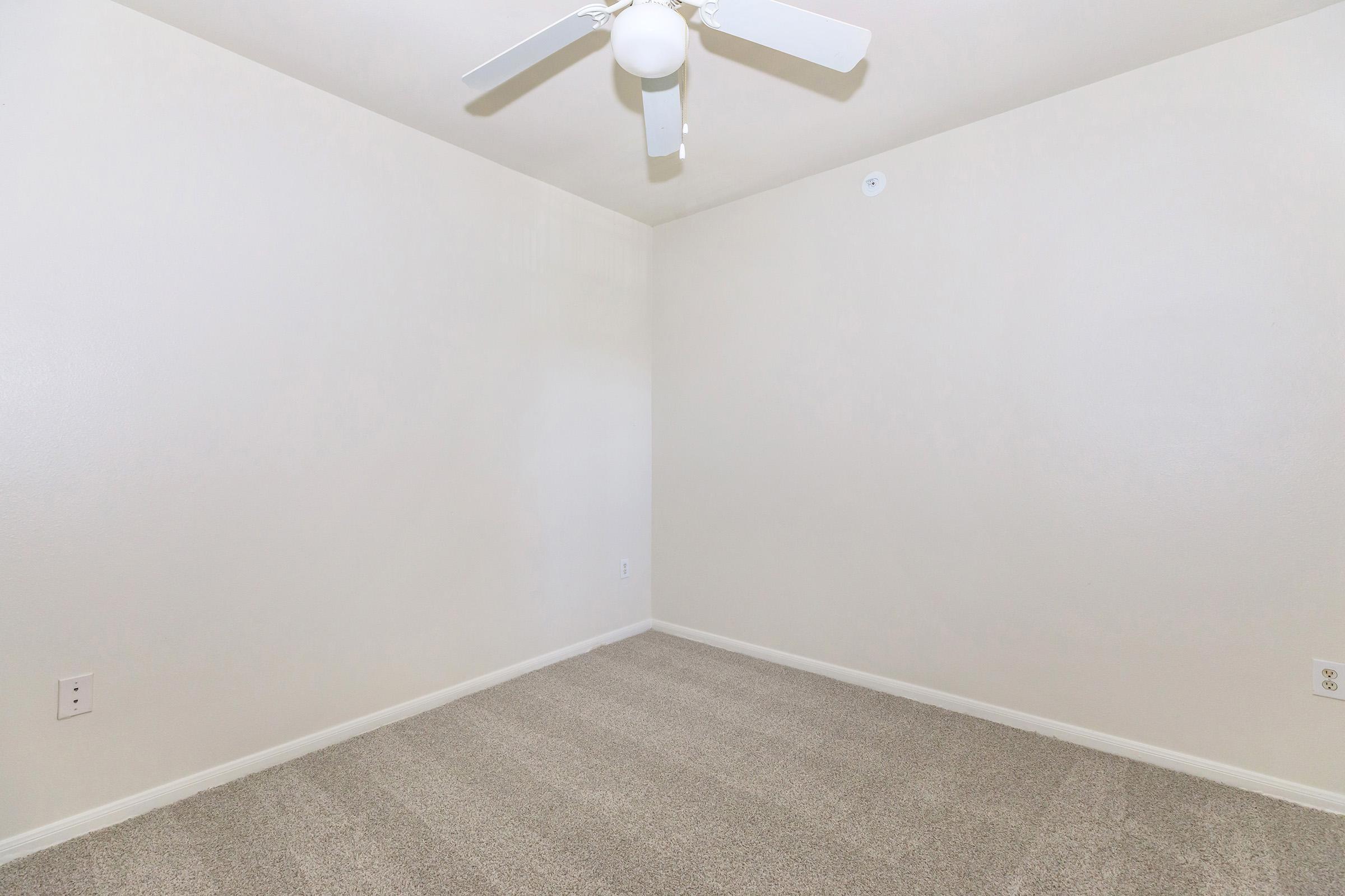 An empty room with light-colored walls and a ceiling fan. The floor is covered with beige carpeting, and there is minimal decor or furniture present. The room appears spacious and well-lit, with a small electrical outlet visible on one wall.