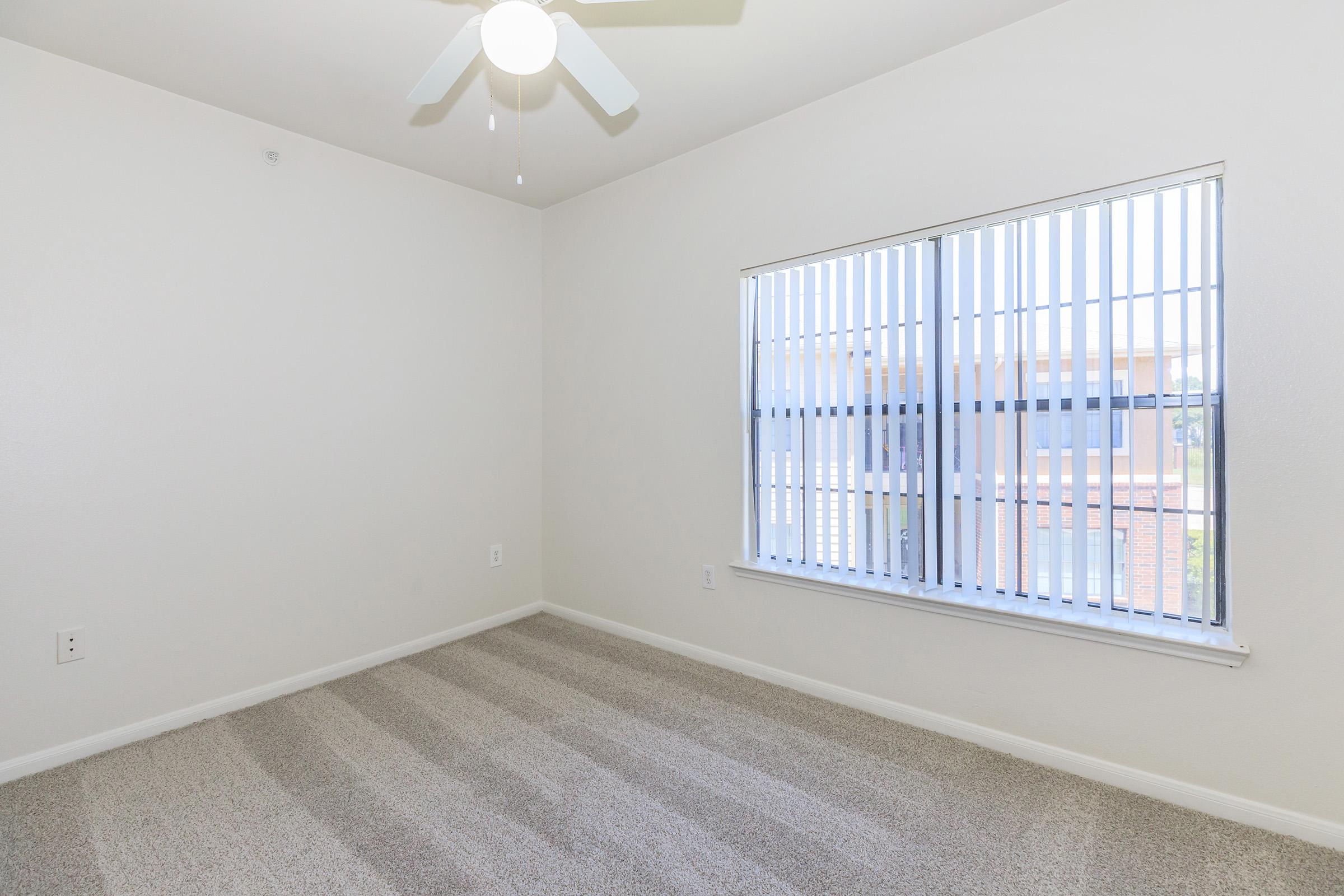 A vacant room with light gray carpet and beige walls. It features a ceiling fan and a large window with horizontal blinds, allowing natural light to enter. The room appears clean and well-maintained, ideal for use as a bedroom or office space.