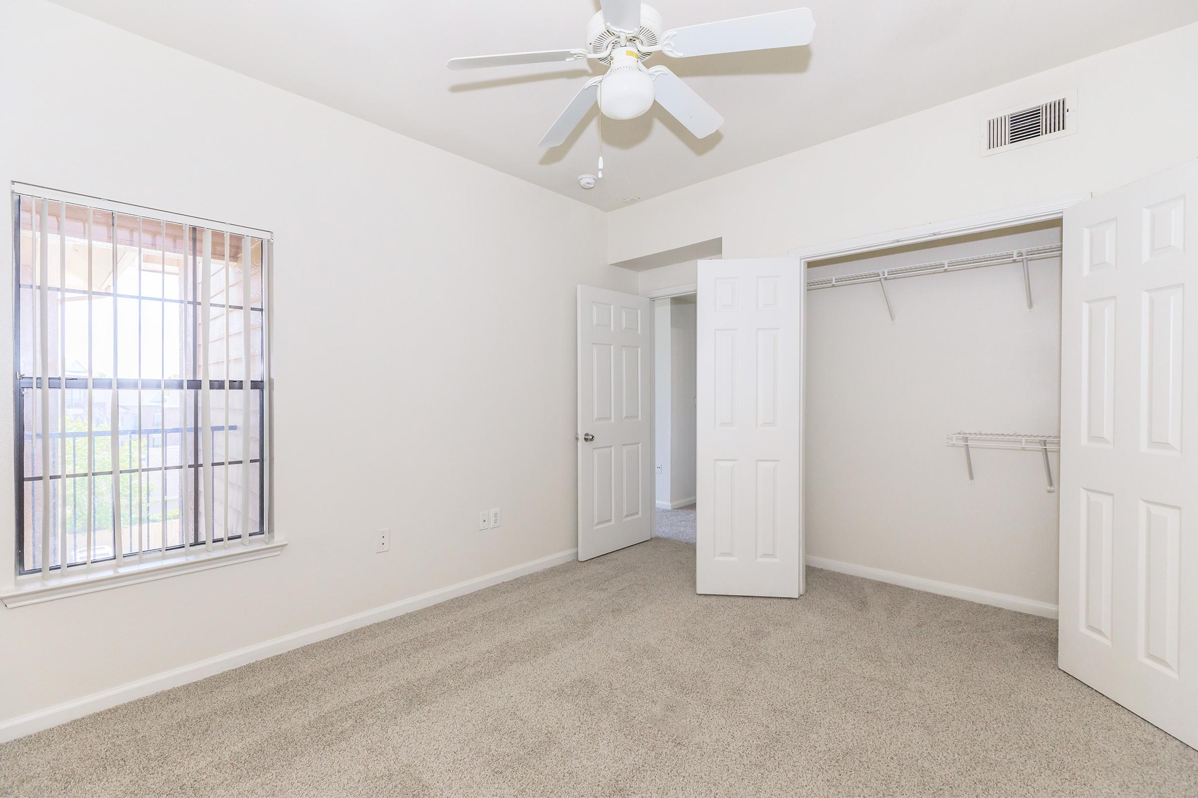 A spacious, vacant bedroom featuring light beige carpet, white walls, and a ceiling fan. The room includes a large window with blinds and a double-door closet with a hanging rod. Natural light illuminates the space, creating a bright and inviting atmosphere.