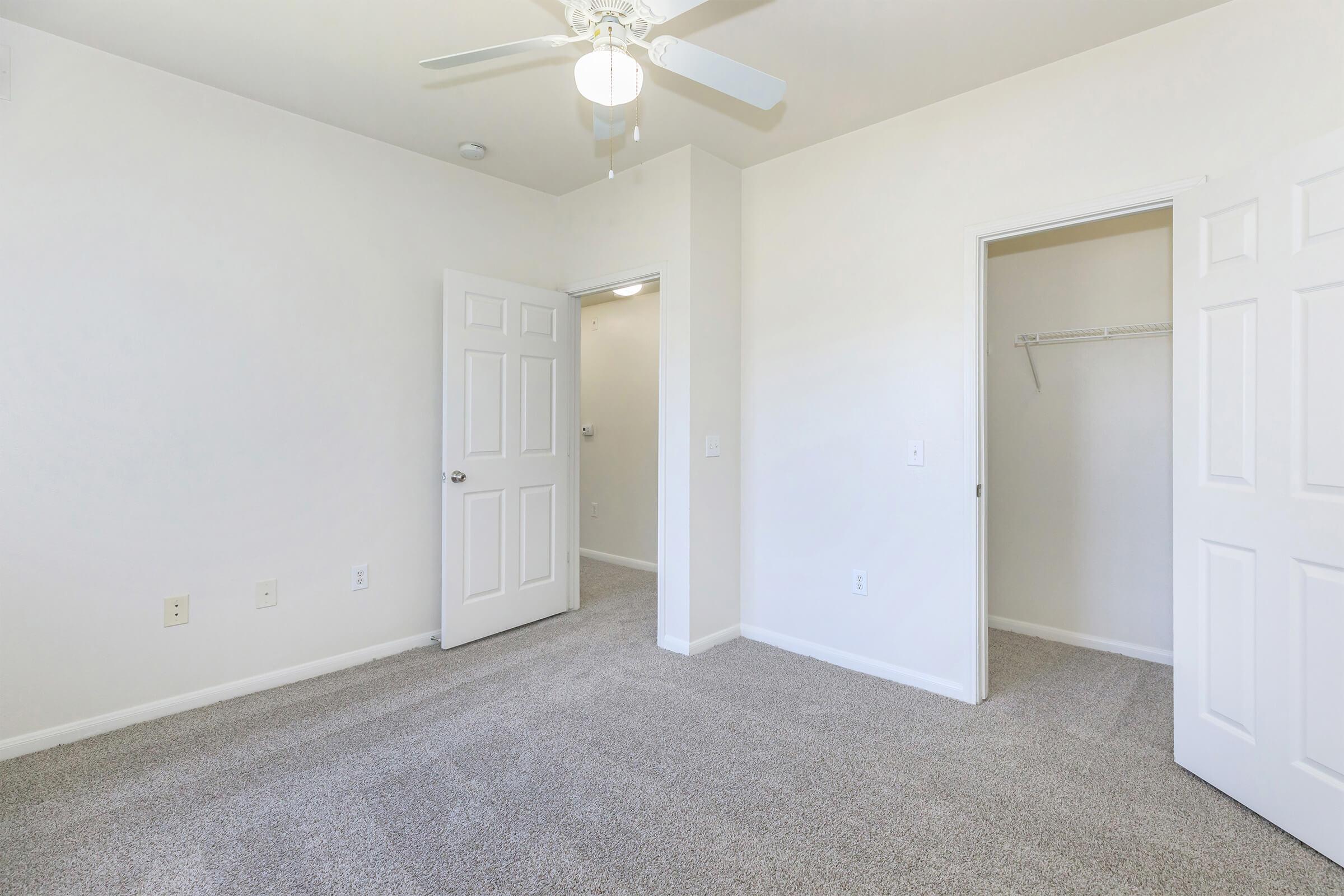 Spacious, empty room with neutral-colored walls, light carpet, and a ceiling fan. Two open doors lead to adjacent spaces, one featuring a built-in closet. Natural light illuminates the area, creating a bright and airy atmosphere. Ideal for personalizing as a bedroom or office.