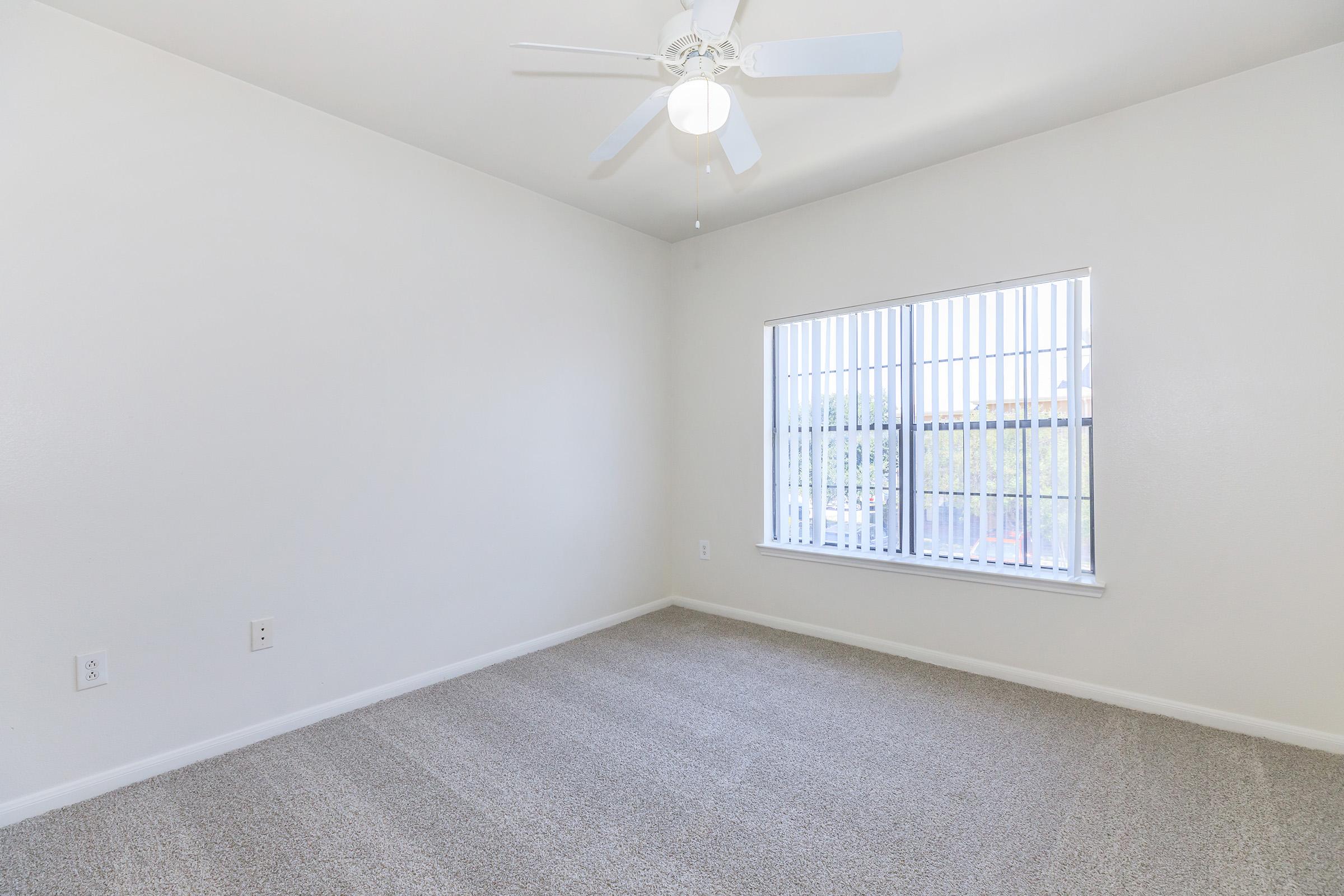Empty room with light-colored walls and a ceiling fan. The floor is covered with beige carpet. There's a window with vertical blinds allowing natural light to enter, and the overall atmosphere is bright and spacious.