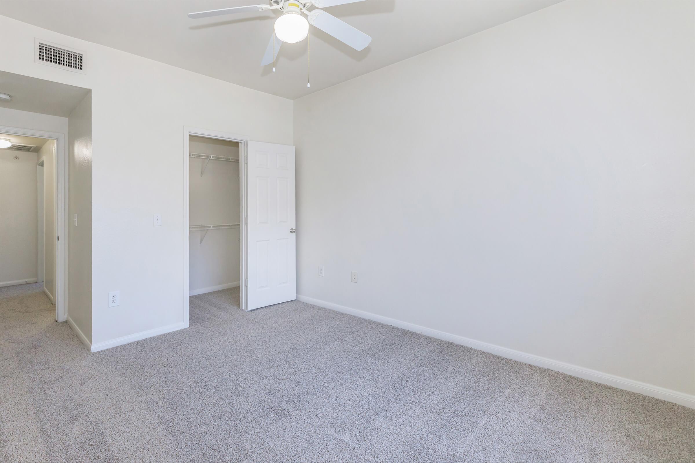 A vacant room with beige carpet flooring and white walls. The room features a ceiling fan, a closed door leading to a closet, and an entryway partially visible on the left. The space is well-lit and minimalistic, offering an empty canvas for decoration and furnishing.