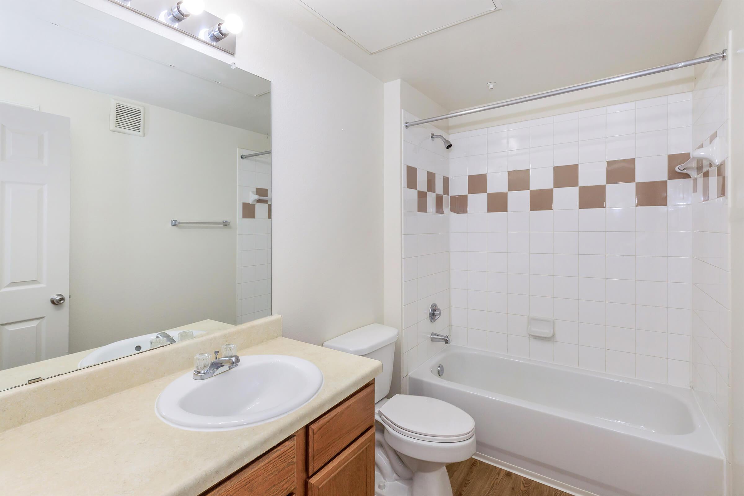 A clean, modern bathroom featuring a bathtub with a shower, a white tiled wall with brown accents, a large mirror above a sink with wooden cabinetry, and a white toilet. Natural light illuminates the space, highlighting the neutral color scheme and spacious layout.