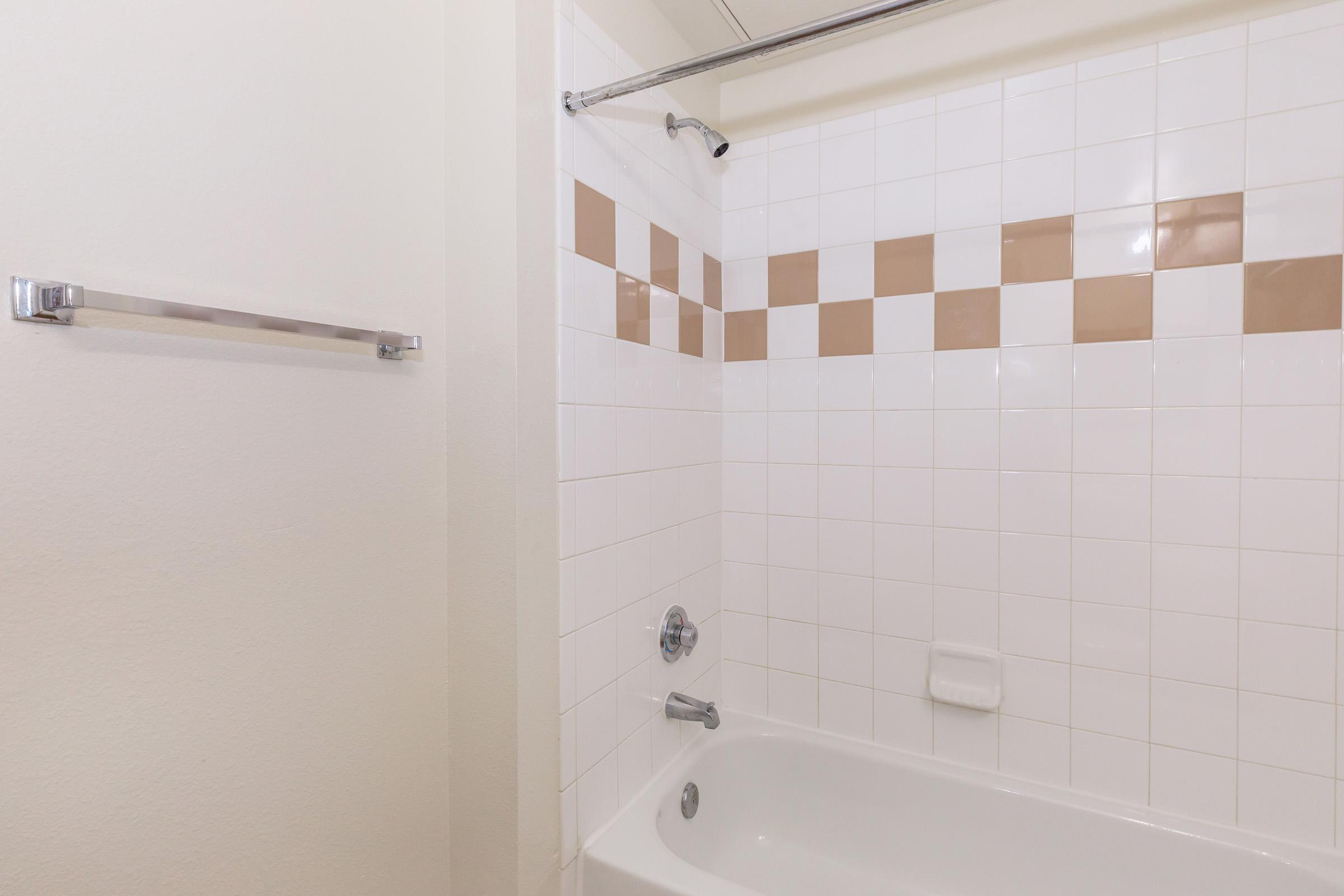 A clean bathroom featuring a white bathtub with a chrome faucet, white tiles, and beige decorative tiles along the wall. A shower curtain rod is installed above the tub, and there is a towel rack on the left wall. The overall decor is simple and airy, providing a fresh and inviting atmosphere.
