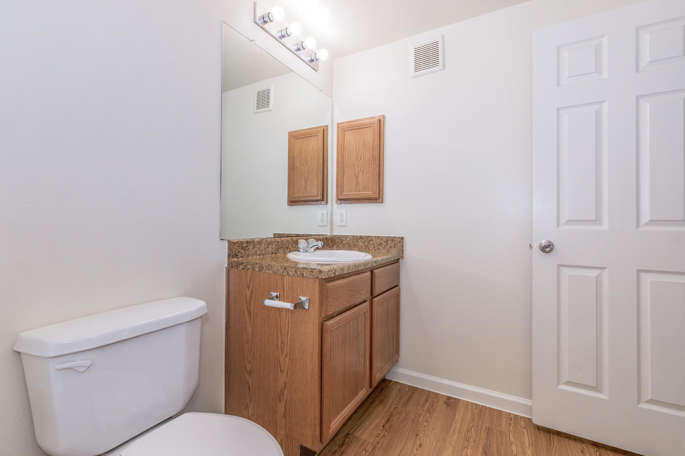 A clean bathroom featuring a white toilet, a wooden vanity with a sink, and a large mirror above the sink. The walls are painted light colors, and there are two wooden cabinets above the sink. The floor has a wood-like appearance, and a door is partially visible on the right side.