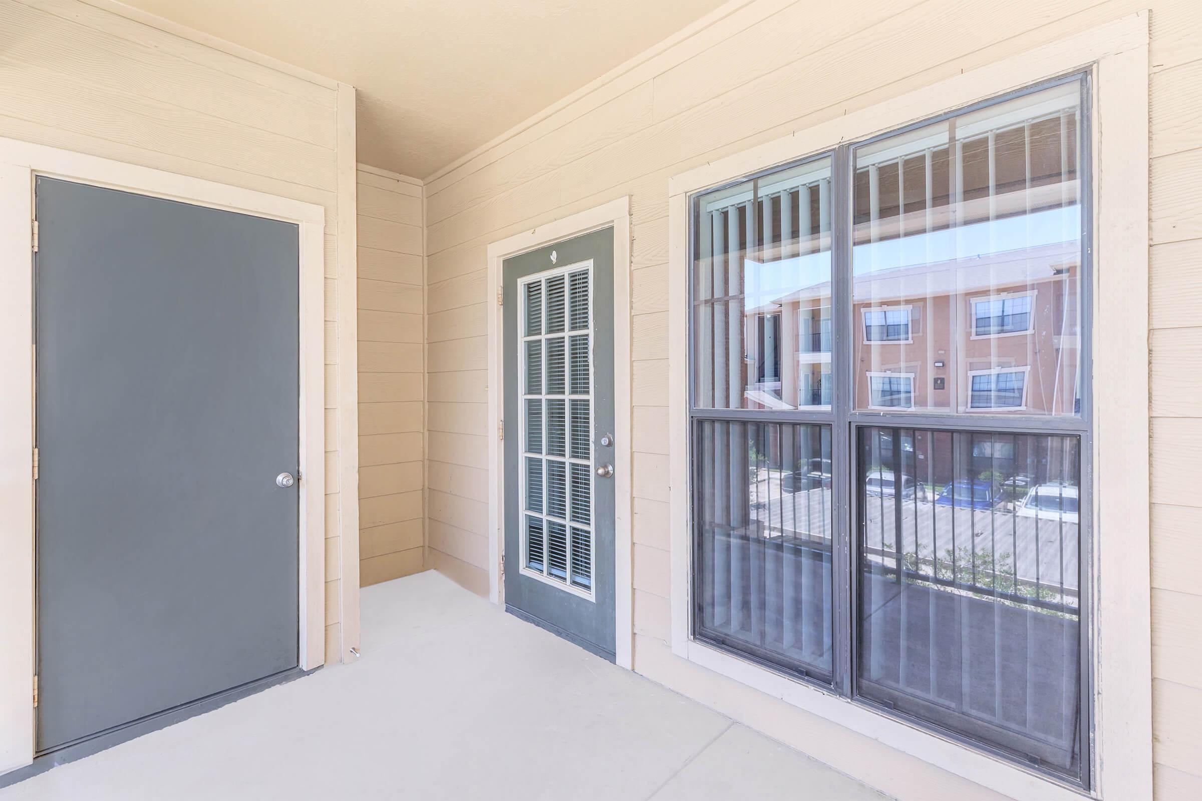 A view of a covered entryway featuring two doors—one gray door on the left and a glass-paned door on the right. Large windows beside the doors offer a view of the exterior surroundings, including nearby buildings. The walls are light-colored with a smooth surface, and the floor is neutral.