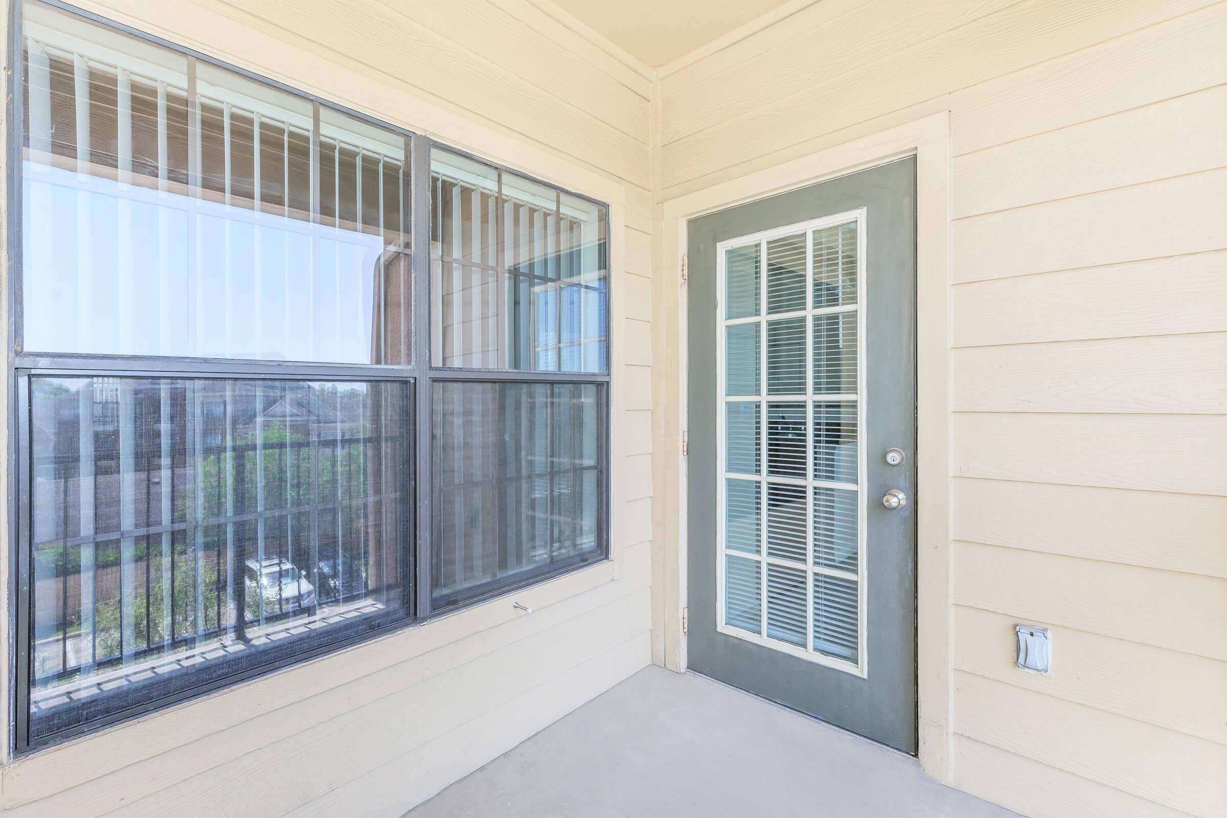 A corner balcony area featuring a window with grids, a door with glass panels, and light-colored wood siding. The view extends outside, showcasing greenery and structures in the distance. The space has a clean, modern appearance with natural light flooding in.