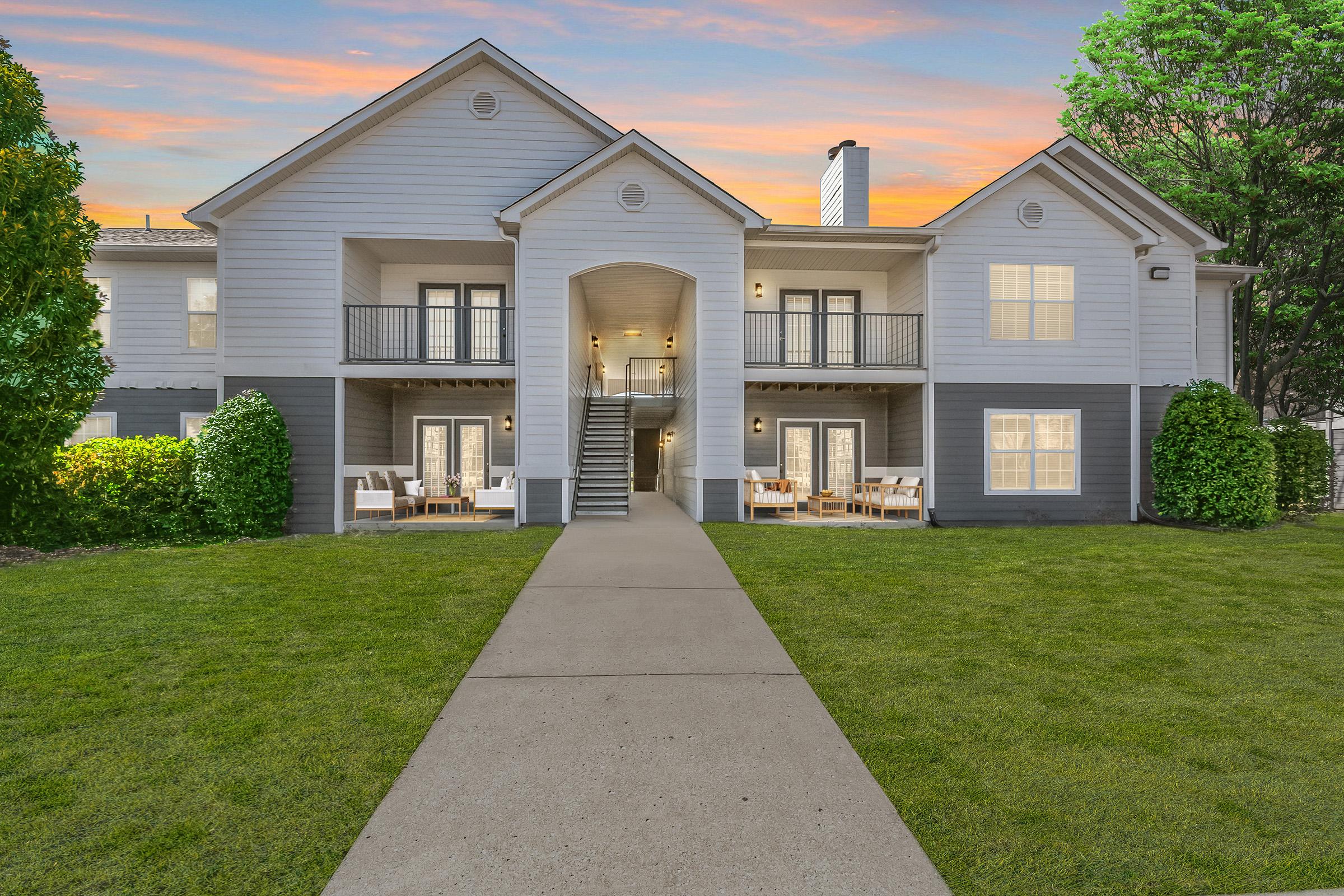 A two-story residential building with a modern design, featuring a light gray exterior, balconies, and large windows in Lebanon, TN. There is a landscaped lawn in front with a concrete path leading to the entrance. The sky above shows a colorful sunset, enhancing the inviting atmosphere of the property.