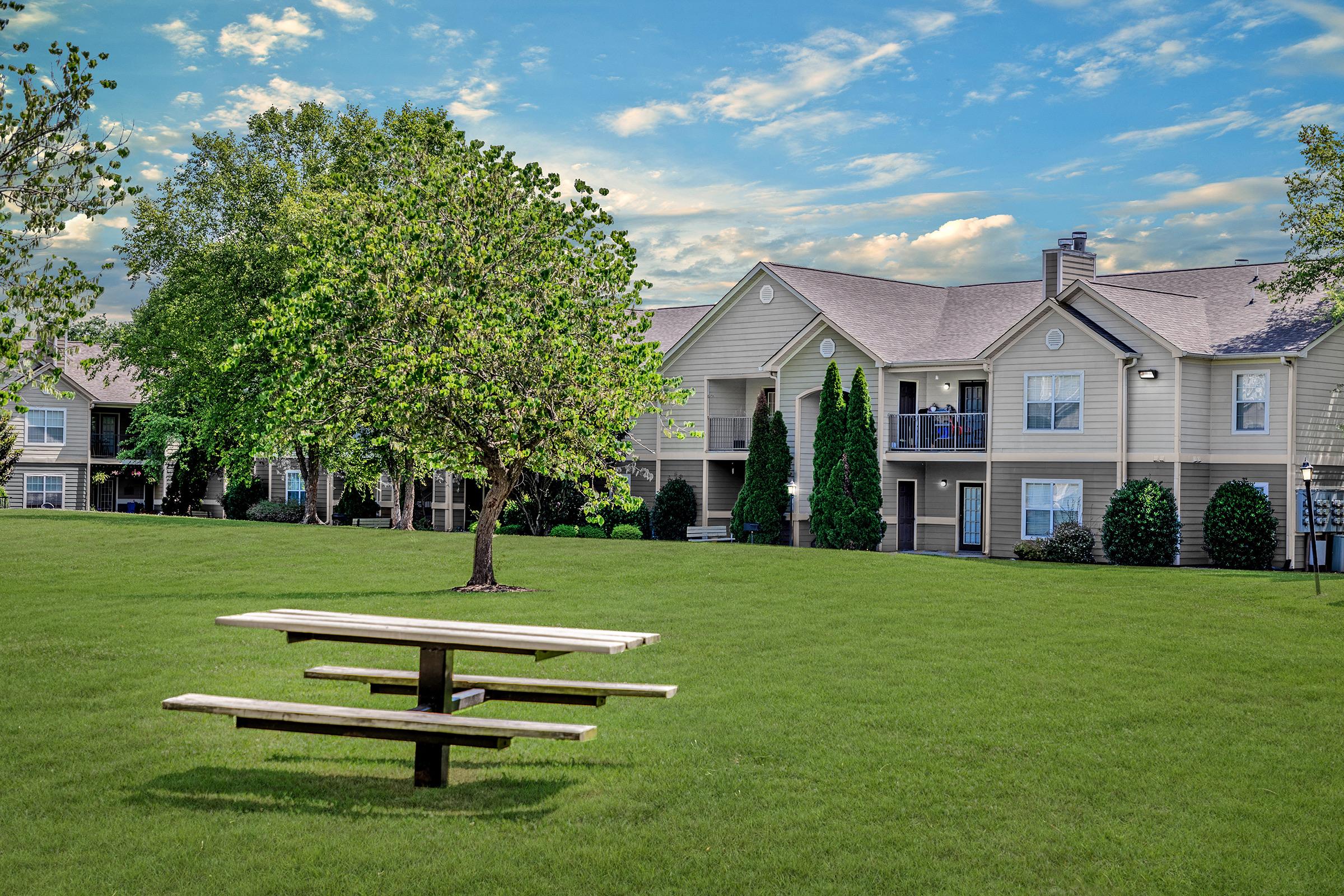 A grassy area with a picnic table in the foreground, surrounded by trees in Lebanon, TN. In the background, there are two-story apartment buildings with balconies, against a bright blue sky with scattered clouds. The scene conveys a peaceful suburban outdoor setting.