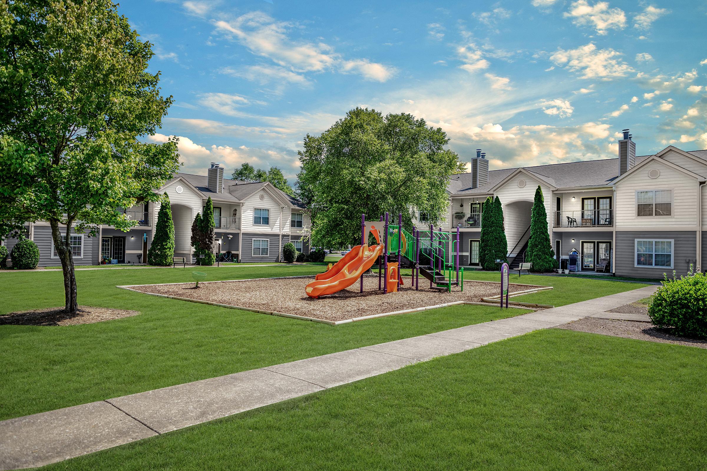 A playground featuring a colorful slide and climbing structure, situated in a grassy area surrounded by residential buildings. The landscape includes well-maintained grass, trees, and walking paths under a partly cloudy sky.