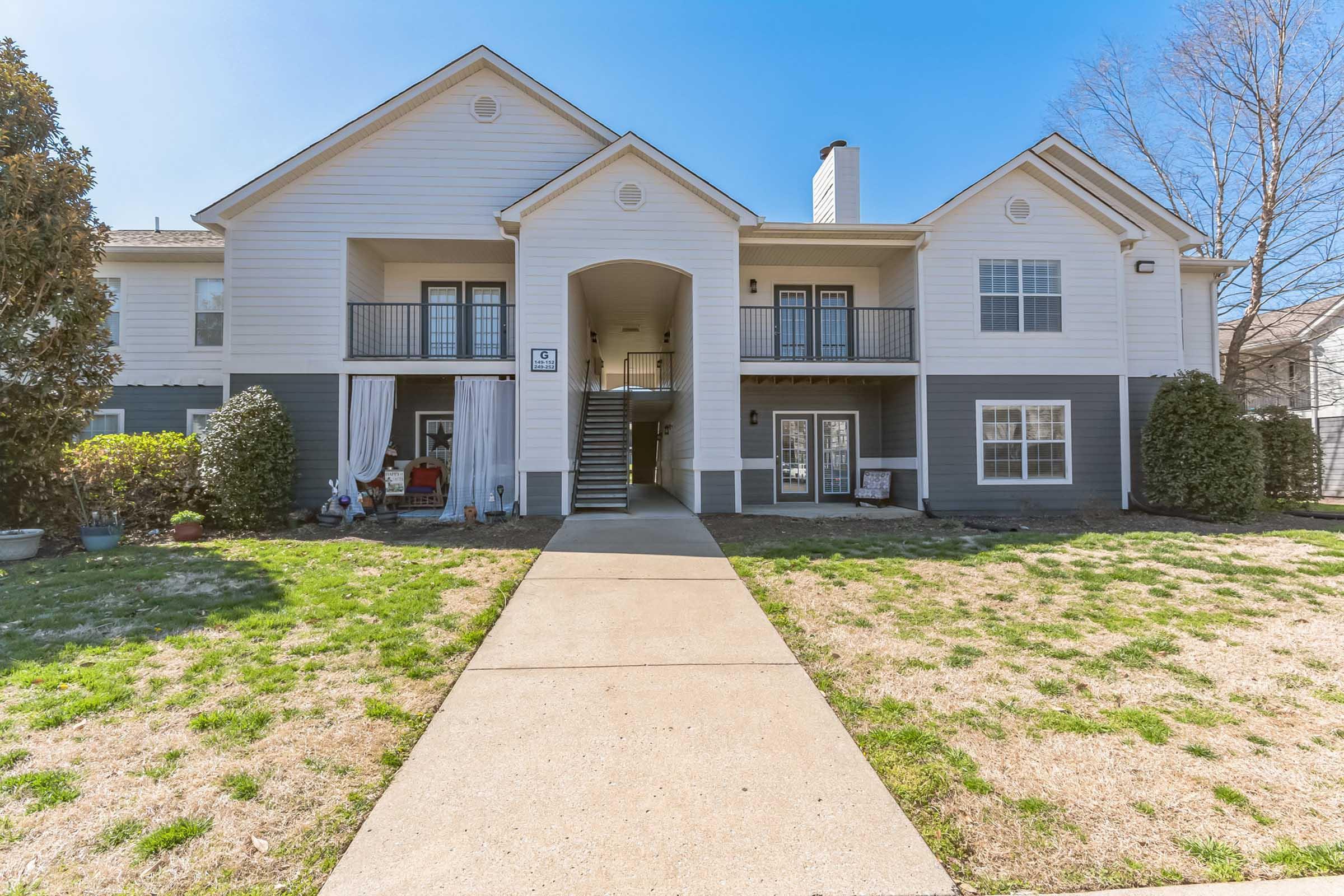 A two-story residential building with a light exterior and dark gray accents. There are balconies on the upper level and a covered entrance on the ground floor. The pathway leading to the entrance is flanked by grass, and there are potted plants near the entrance. Trees are visible in the background.
