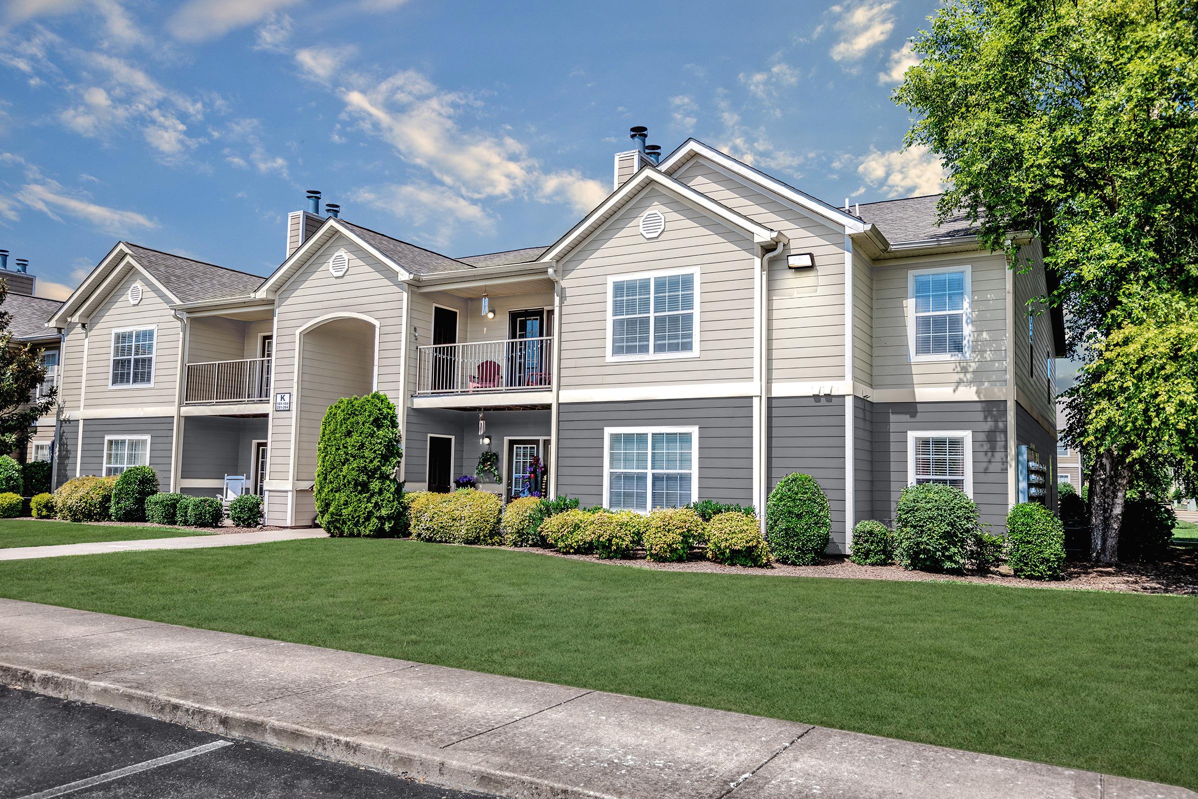 A modern two-story apartment building with a well-maintained exterior. The building features light-colored siding, a welcoming porch with chairs, and neatly trimmed bushes in the yard. A sunny blue sky with scattered clouds completes the inviting scene.