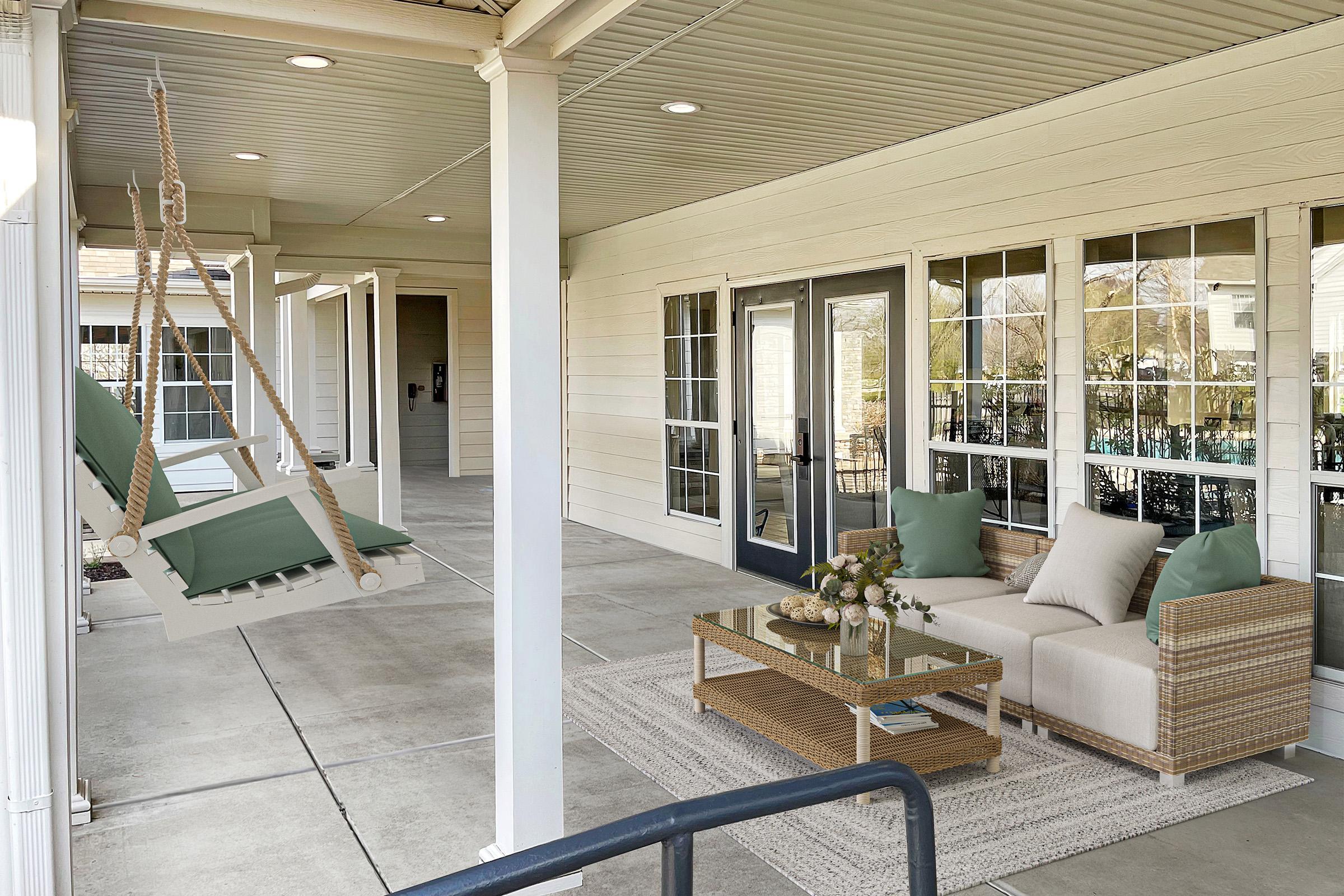A cozy porch with a swing chair and a sectional sofa with green cushions. A coffee table sits on a light rug, surrounded by large windows that let in natural light. The space is designed for relaxation and socializing, featuring a neutral color palette and a welcoming atmosphere.