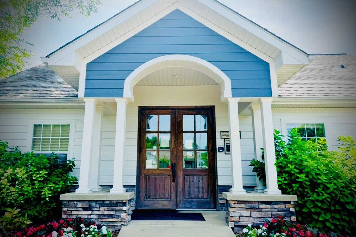 A welcoming entrance to a house featuring a large wooden double door framed by white columns. The home has a blue and white exterior, a neatly landscaped front with green shrubs and colorful flowers, and large windows that enhance its inviting appearance.