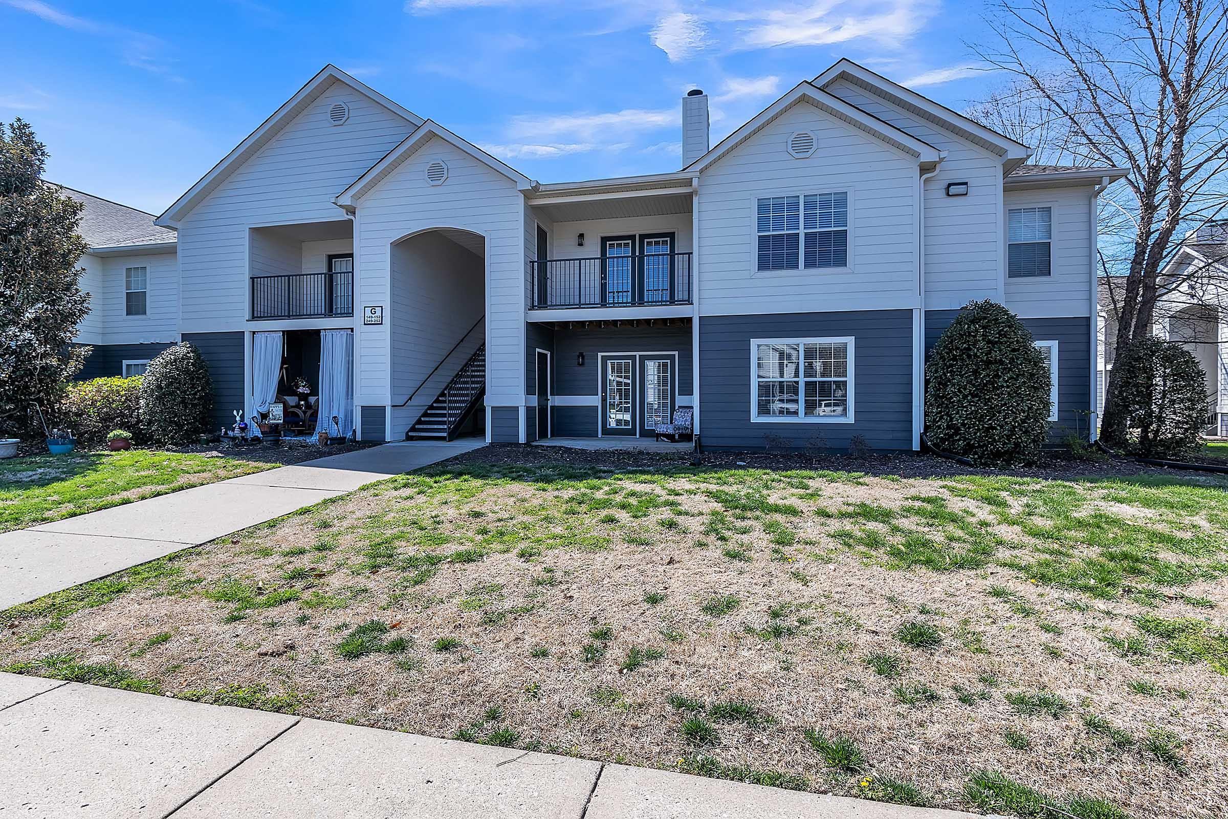 Two-story apartment building with a light-colored façade and dark blue accents, Lebanon, Tennessee. The entrance features a covered porch with stairs leading up. Lush green grass and shrubs surround the building, with a clear blue sky overhead.