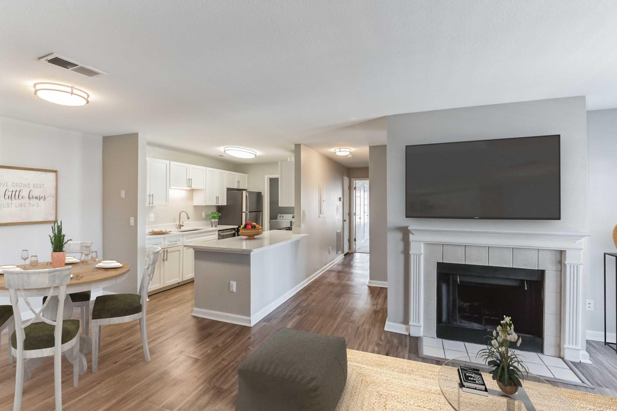 A modern living room and kitchen area featuring a cozy fireplace, a flat-screen TV mounted on the wall, a dining table set for four, and white cabinetry in the kitchen. The space is bright with neutral colors and stylish decor, creating a welcoming and comfortable atmosphere.