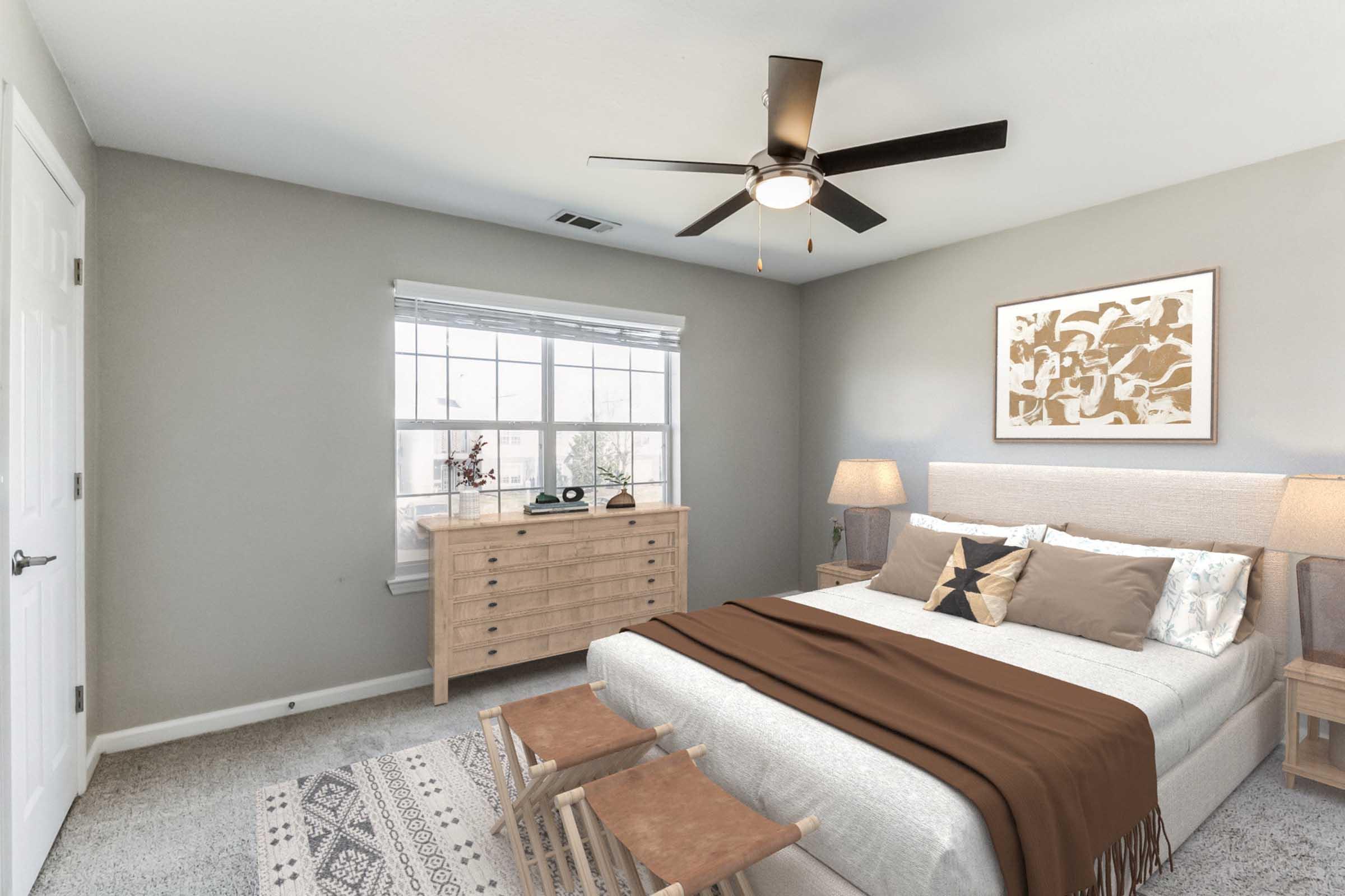 A modern bedroom featuring a queen-sized bed with neutral bedding and a brown throw, two wooden stools at the foot of the bed, a light wood dresser, bedside tables with lamps, and a ceiling fan. A large window lets in natural light, and minimalist decor adorns the walls.