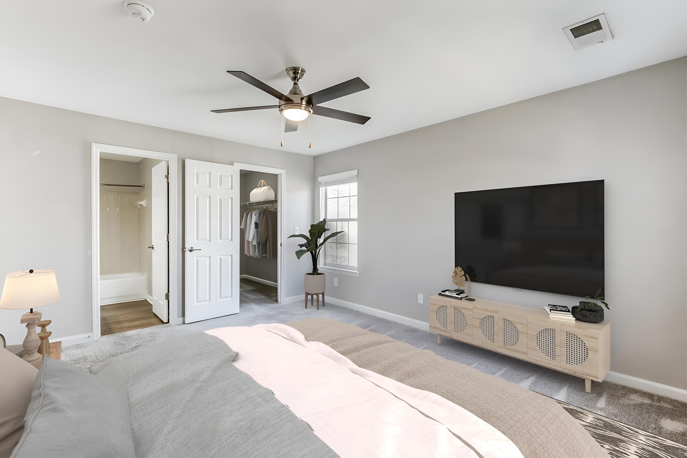 A modern bedroom featuring a light gray color scheme, a comfortable bed with layered bedding, a TV mounted on the wall, and a ceiling fan. There are double doors leading to a bathroom and a closet area. A small plant and a lamp add a touch of warmth to the space.