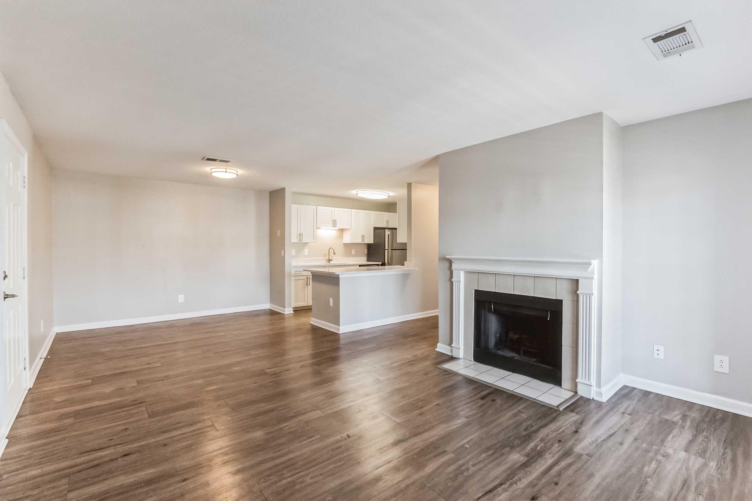 A spacious living room featuring hardwood floors, a fireplace, and bathed in natural light. The room connects to a modern kitchen with white cabinets and an island. The neutral color palette creates a warm and inviting atmosphere. Suitable for comfortable living and entertaining.