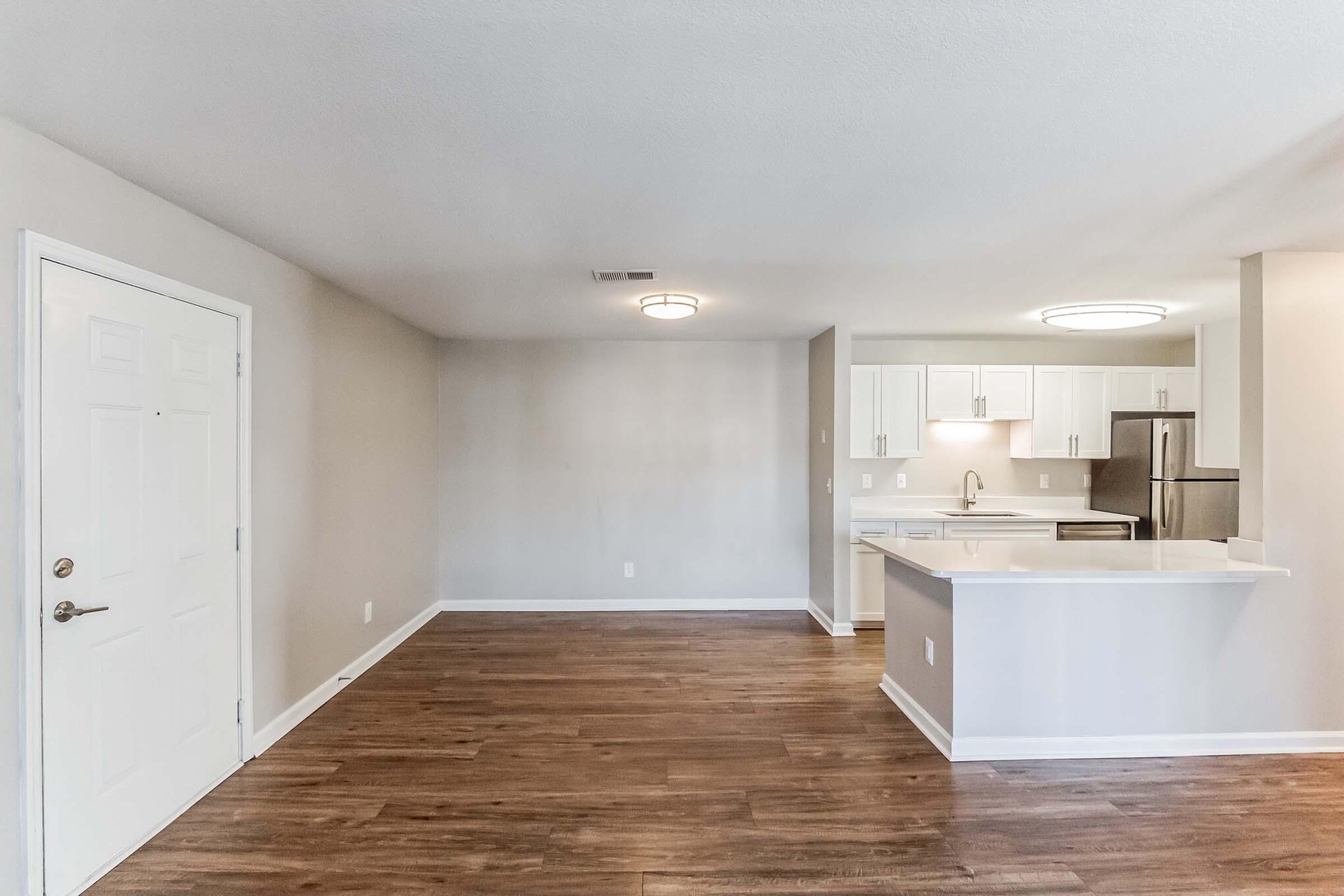 Spacious open-concept living area with hardwood flooring, featuring a kitchen in the background equipped with modern appliances, white cabinetry, and a breakfast bar. Walls are painted in neutral tones, and ample natural light fills the space, creating a warm and inviting atmosphere.