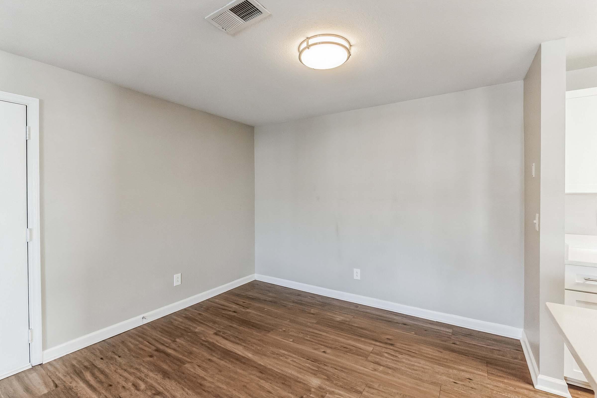 A spacious, empty room with light gray walls and a wooden floor. There is a ceiling light fixture and a door on the left side. The right side features a kitchen area with minimal furnishings, creating an open and modern atmosphere. The room is well-lit and ready for decoration.