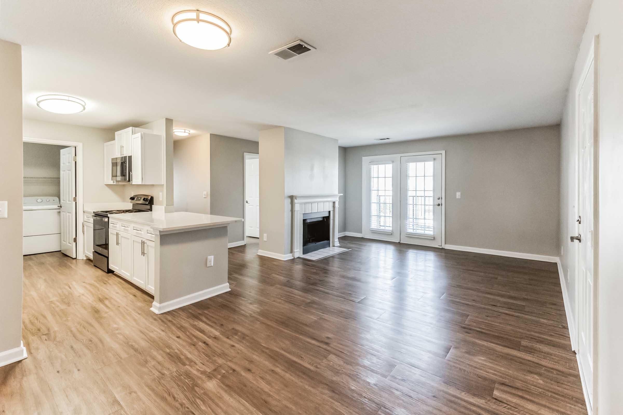 A spacious, modern living area with hardwood floors, a fireplace, and large windows allowing natural light. The kitchen features white cabinets and stainless steel appliances. An adjoining laundry area is visible, and the room is painted in neutral tones, enhancing the open concept layout.