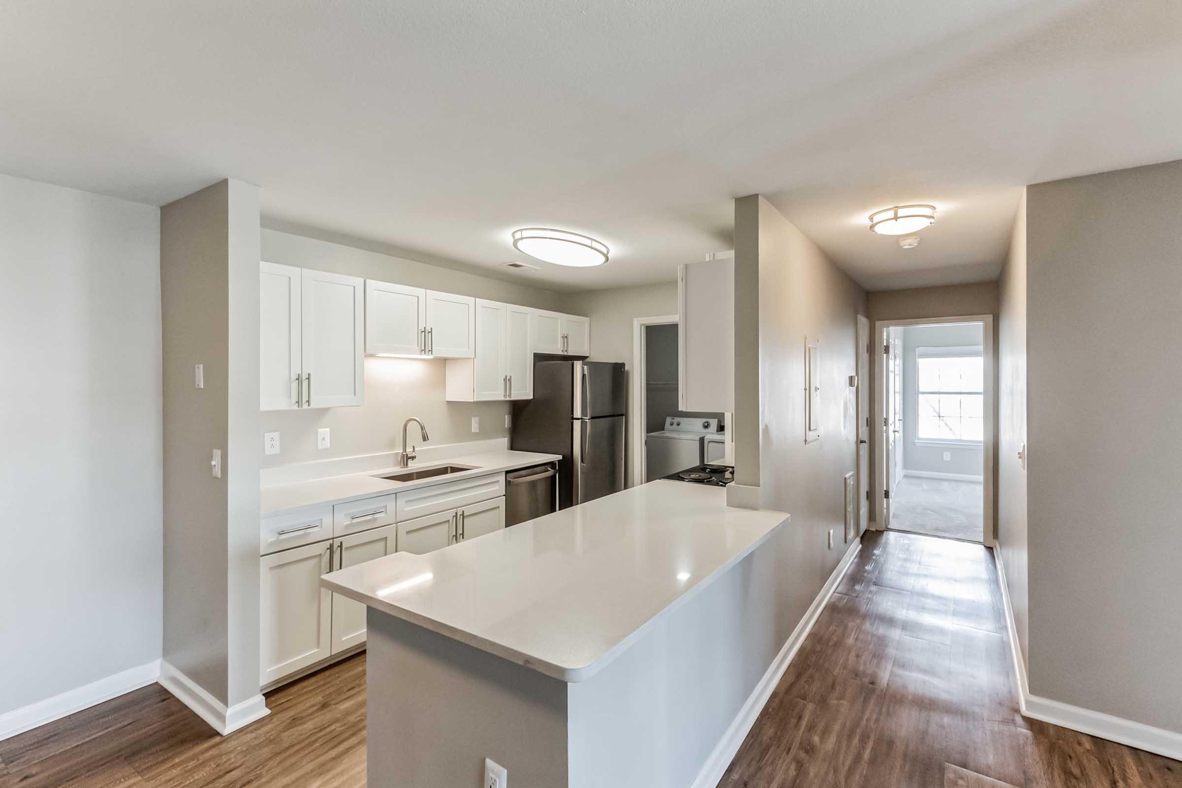 Modern kitchen featuring white cabinetry, stainless steel appliances, and a large island with a light-colored countertop. The space has neutral walls and hardwood flooring, leading to a hallway with natural light visible in the distance.