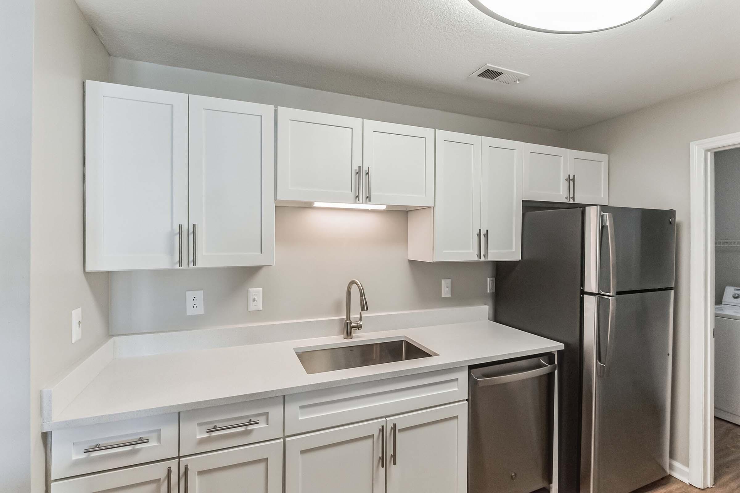 Modern kitchen featuring white cabinets, a stainless steel refrigerator, a double sink with a sleek faucet, and white countertops. Soft lighting illuminates the space, which has light gray walls and wooden flooring. A doorway leads to an adjoining laundry area.