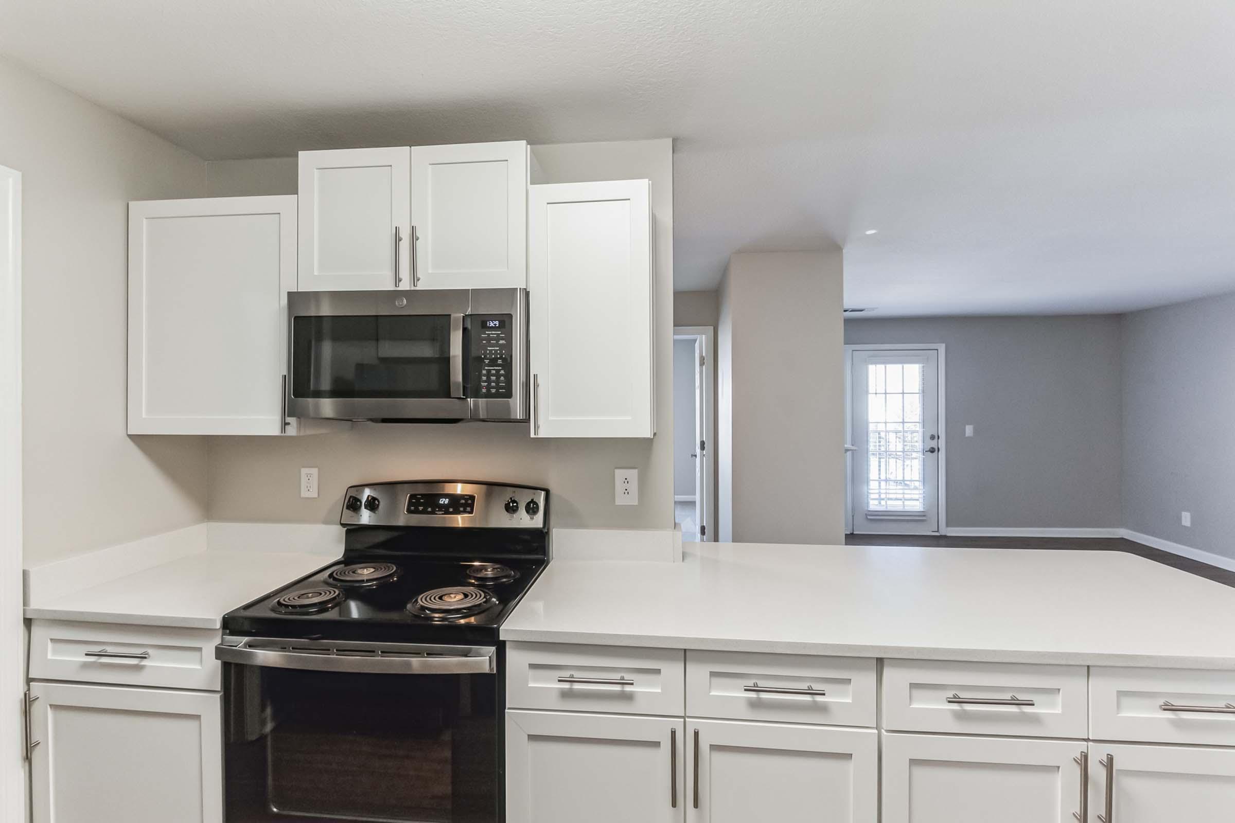 A modern kitchen featuring a white countertop, stainless steel stove, and microwave. The cabinets are white with sleek handles, and there is a spacious layout that connects to a well-lit living area in the background. Large windows provide natural light, enhancing the open feel of the space.