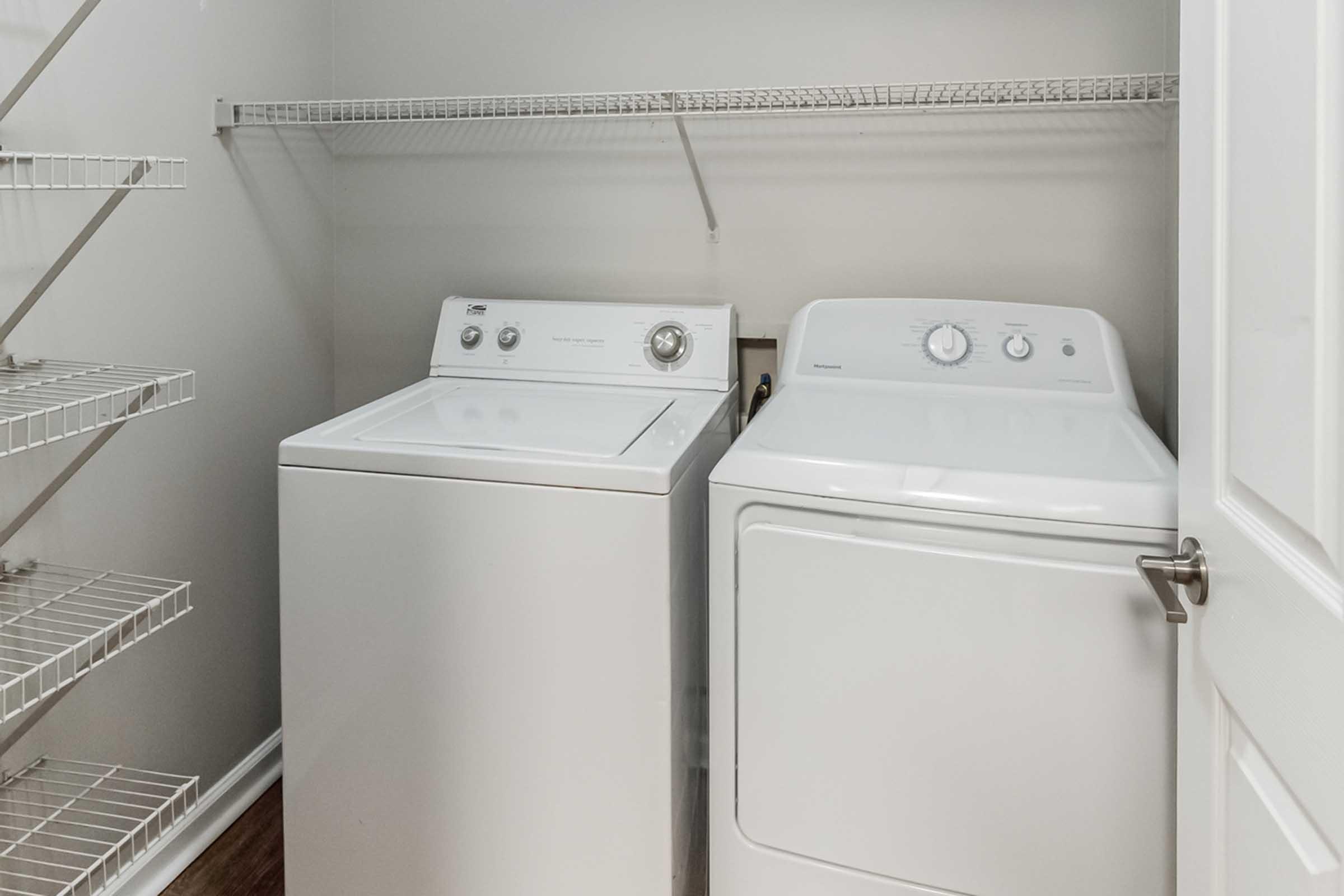 A laundry room featuring a white washing machine and a white dryer placed side by side. There are metal wire shelves on the left for storage, and a door is partially visible on the right, adding to the organized and practical design of the space.