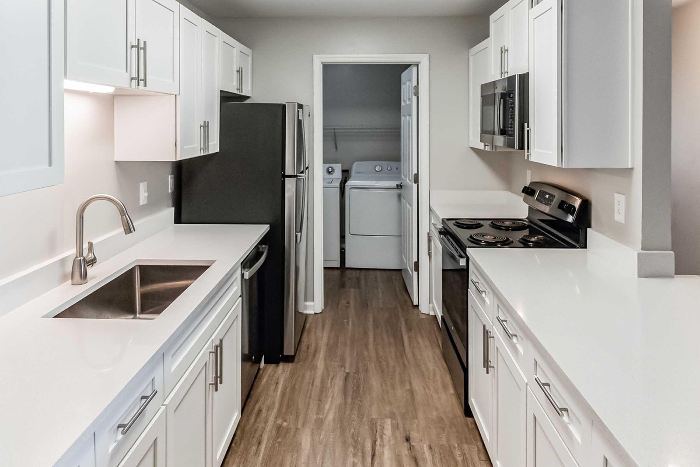 Modern kitchen with white cabinets, stainless steel appliances, and a sleek countertop. Features a double sink, stove, and refrigerator. A laundry area is visible in the background through an open door. The flooring is light wood, complementing the contemporary design.