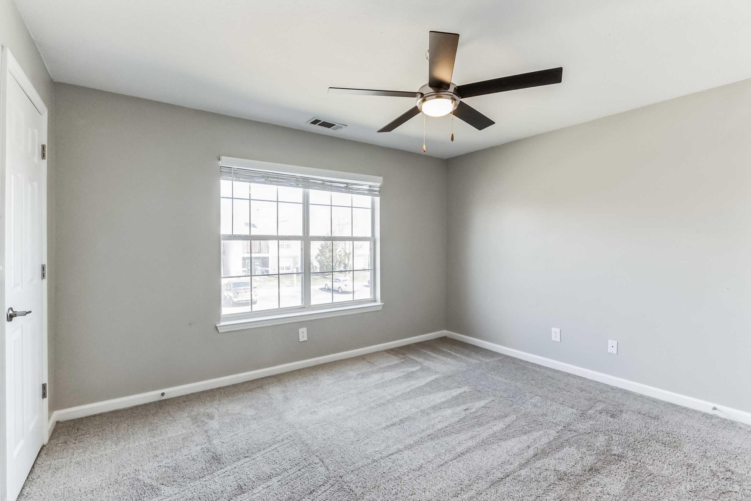 A spacious, empty bedroom with light gray walls and a ceiling fan. The room features a window with multiple panes allowing natural light to enter, and the floor is carpeted in a neutral tone. There are no furnishings, creating a clean and open atmosphere.