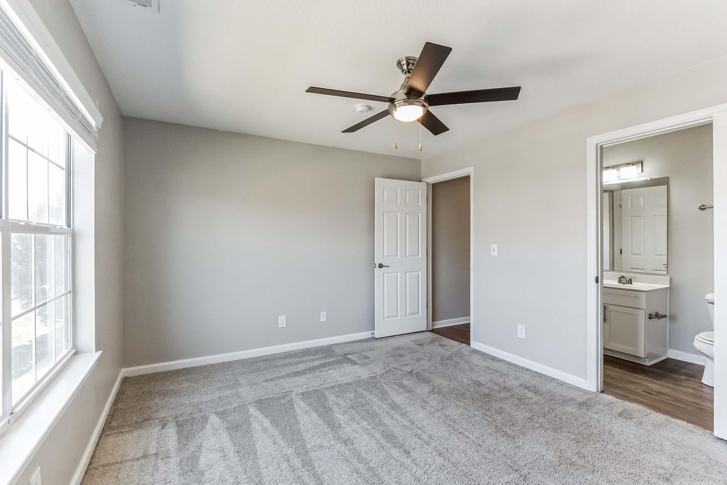 A well-lit, empty room with gray walls and carpet. It features a ceiling fan, a large window letting in natural light, and an open door leading to a bathroom area. The bathroom includes a sink and mirror. The overall feel is clean and spacious, ideal for personal customization.