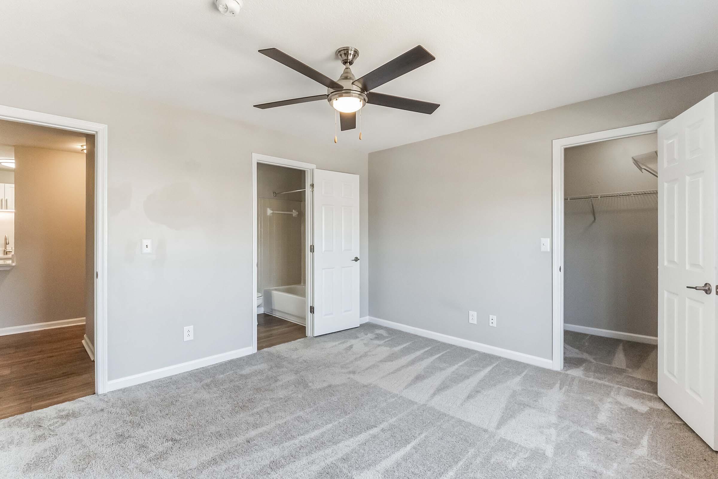 A neutral-colored bedroom featuring light gray walls and plush carpeting. The room has a ceiling fan and an open layout with two doors leading to closets. Natural light illumination is visible, creating a welcoming atmosphere.